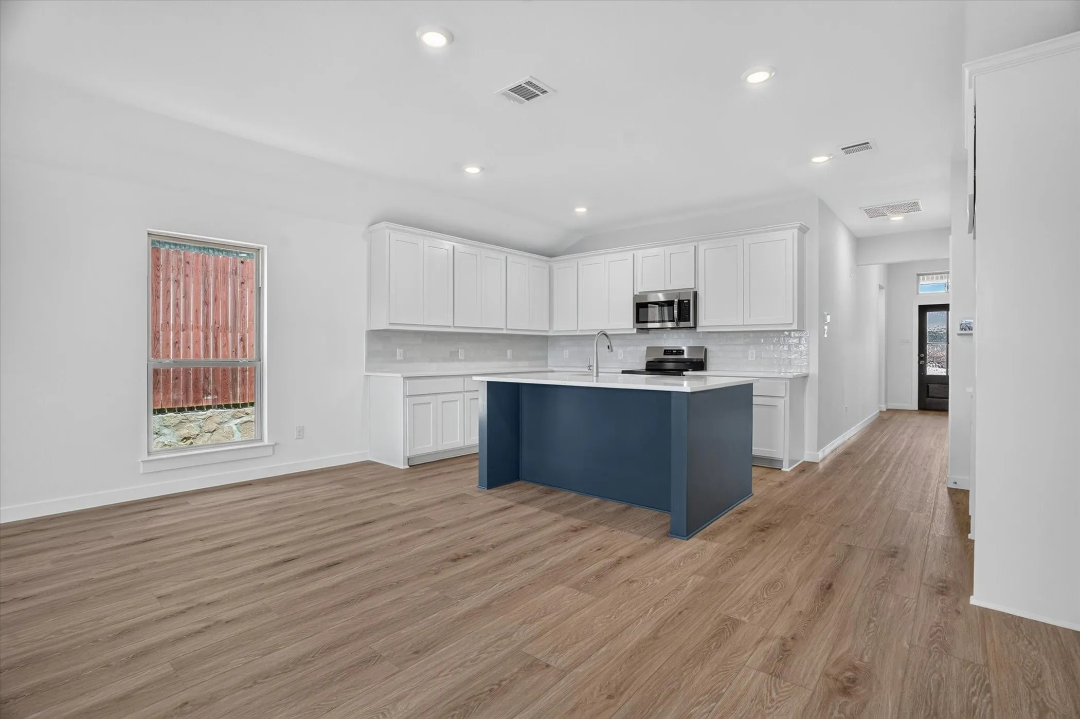 Open-concept kitchen featuring a navy blue island, white shaker cabinetry, stainless steel appliances, white subway tile backsplash, and wood-finish flooring