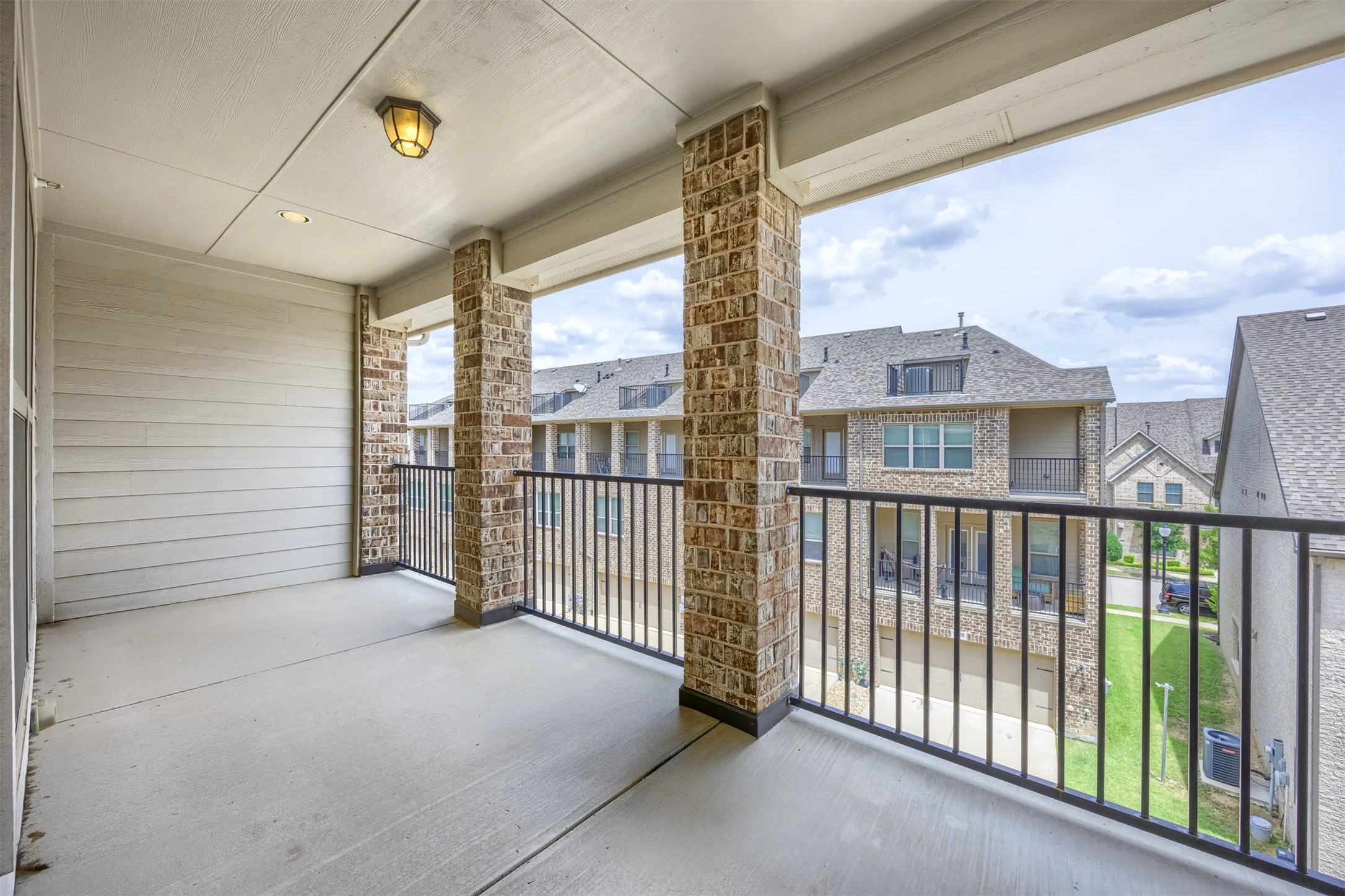 Spacious balcony featuring concrete flooring, brick-clad columns, black metal railing, and a tongue-and-groove ceiling with a surface-mount light fixture