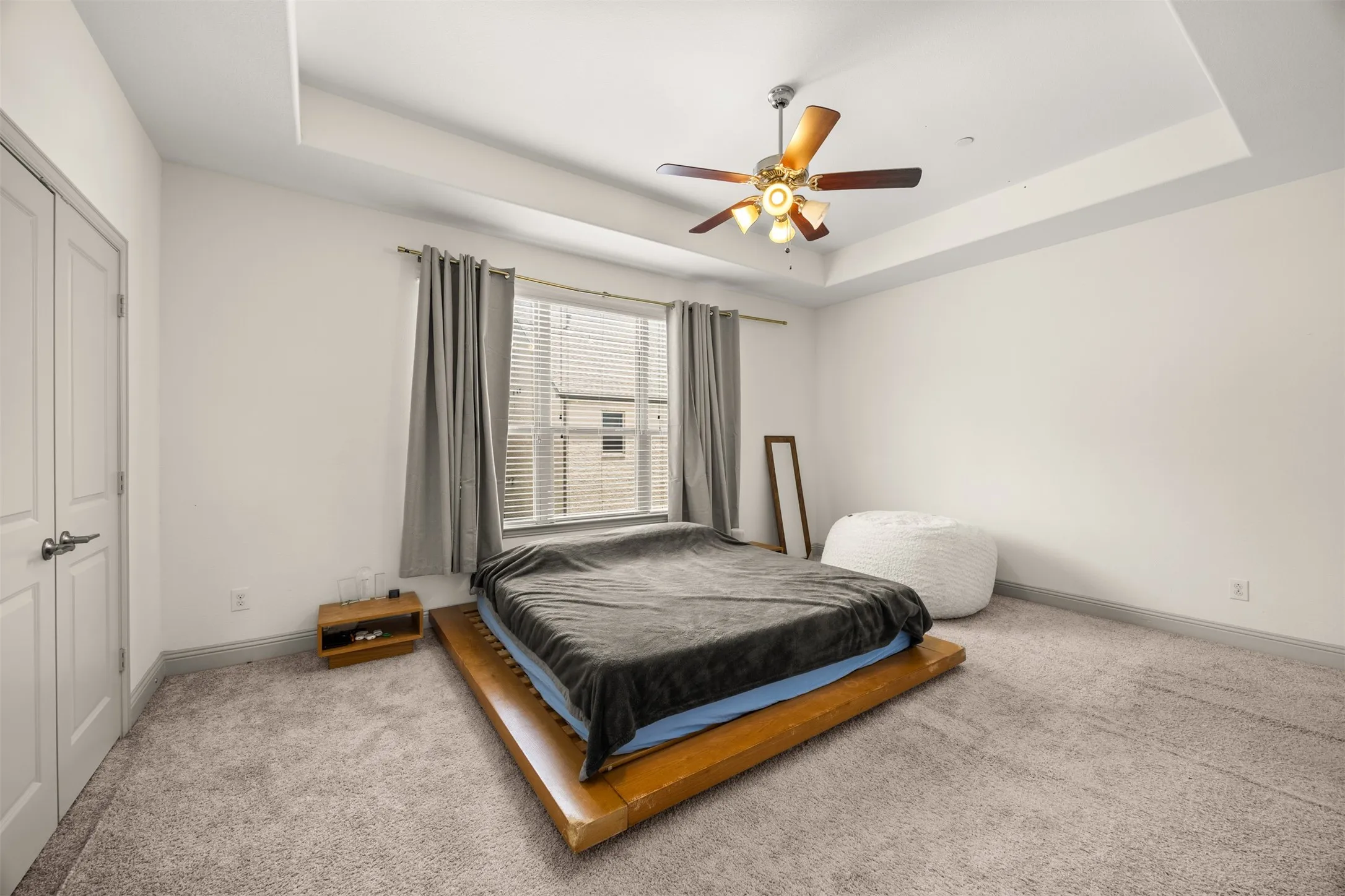Spacious room featuring a tray ceiling with a ceiling fan, light gray carpeting, and a large window with blinds and gray curtains