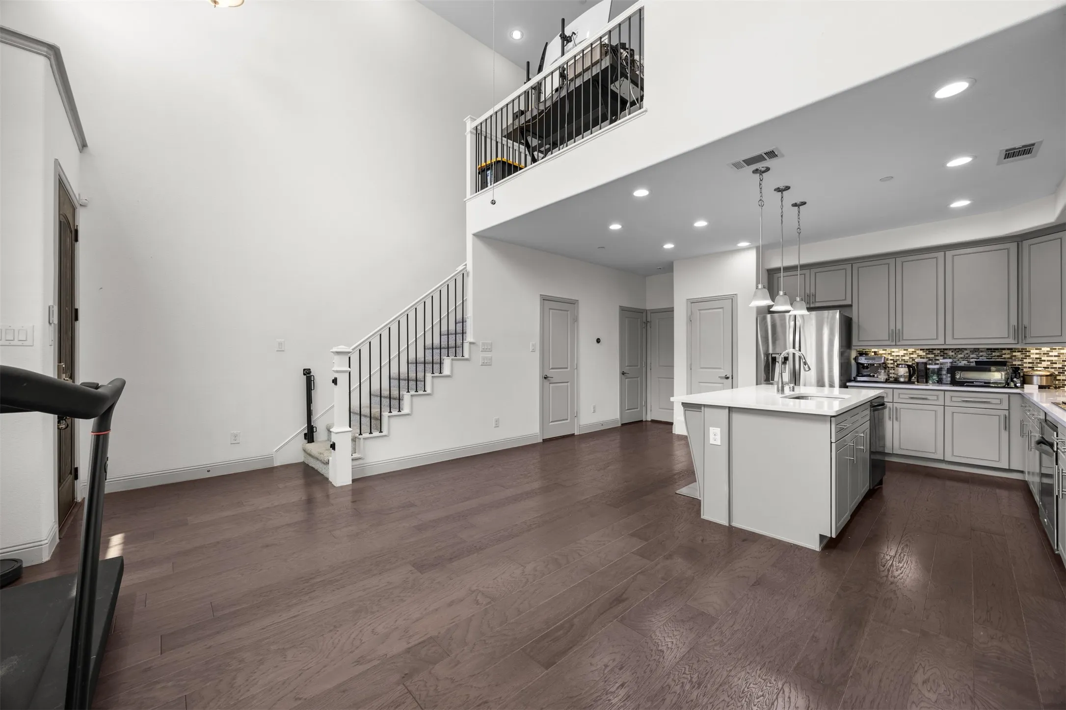 Open-concept living space featuring high ceilings, wood-finish flooring, a contemporary kitchen with a central island and pendant lighting, ample cabinetry, and a staircase leading to a mezzanine
