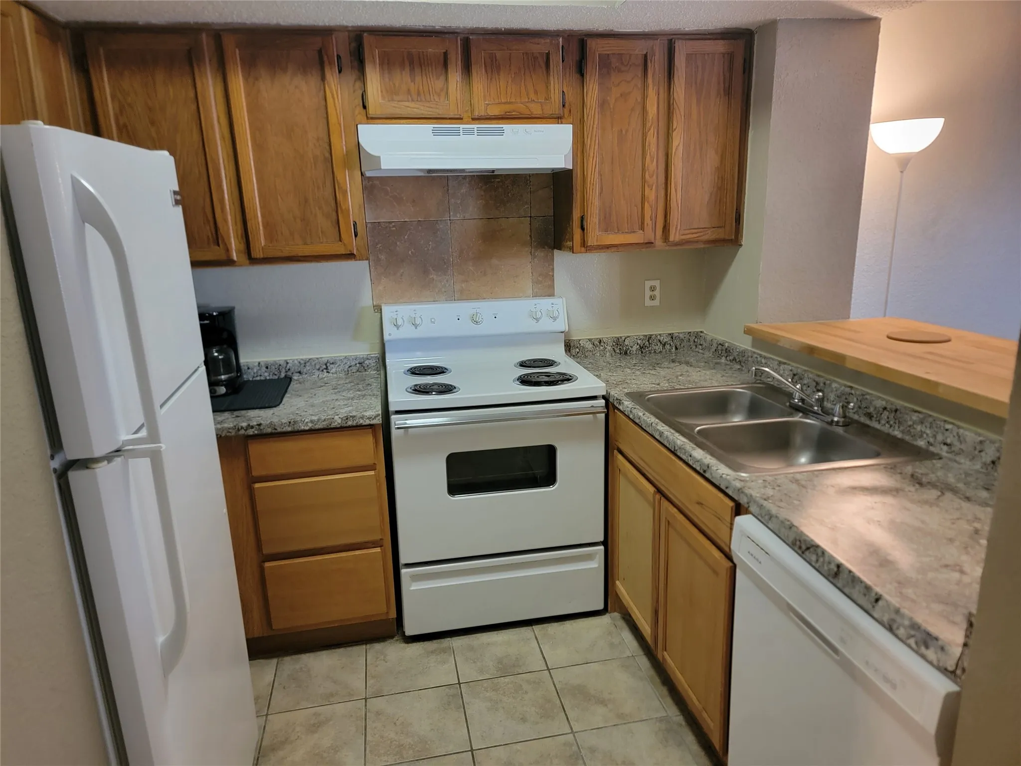 Compact kitchen featuring wood cabinetry, speckled countertops, a double basin stainless steel sink, white range, and tiled flooring