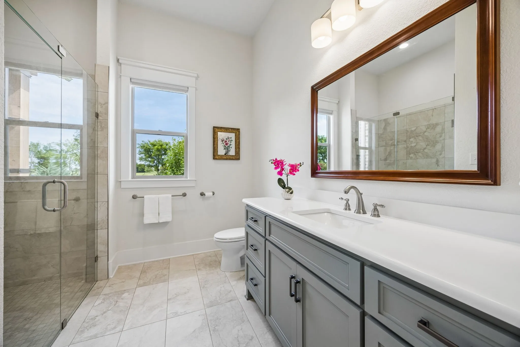 Bathroom featuring a glass-enclosed shower with tile surround, a single vanity with a white countertop and gray cabinetry, a large framed mirror, and light-toned tile flooring