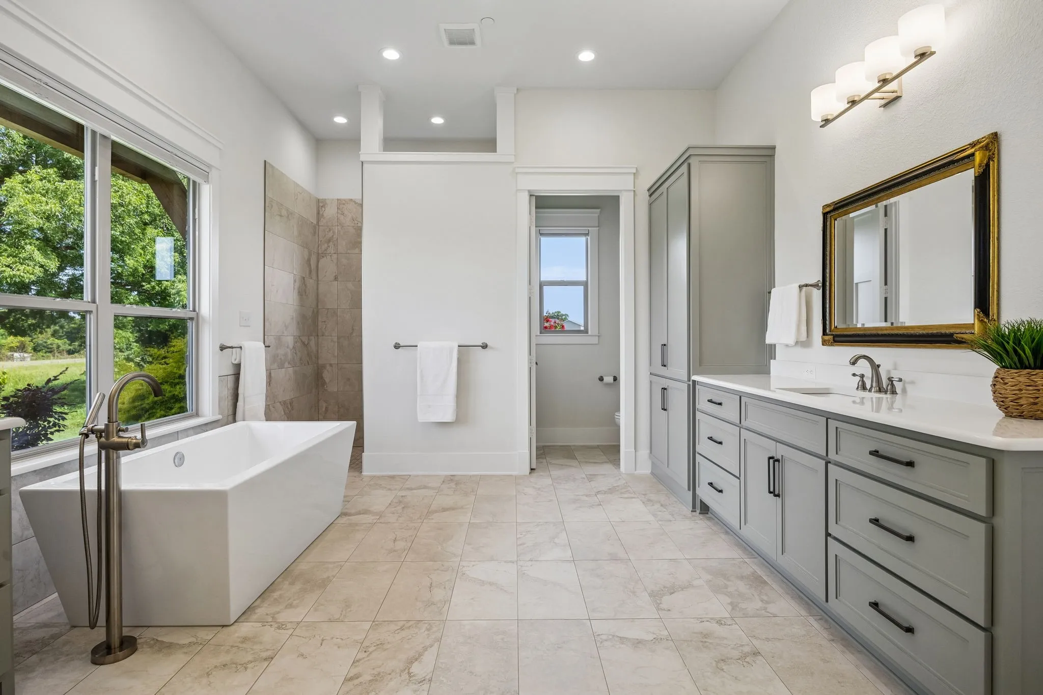The luxurious primary spa bath features large picture windows, heated tile floors, freestanding soaking tub, large roll-in shower, separate vanities with shaker cabinetry and quartz countertops.
