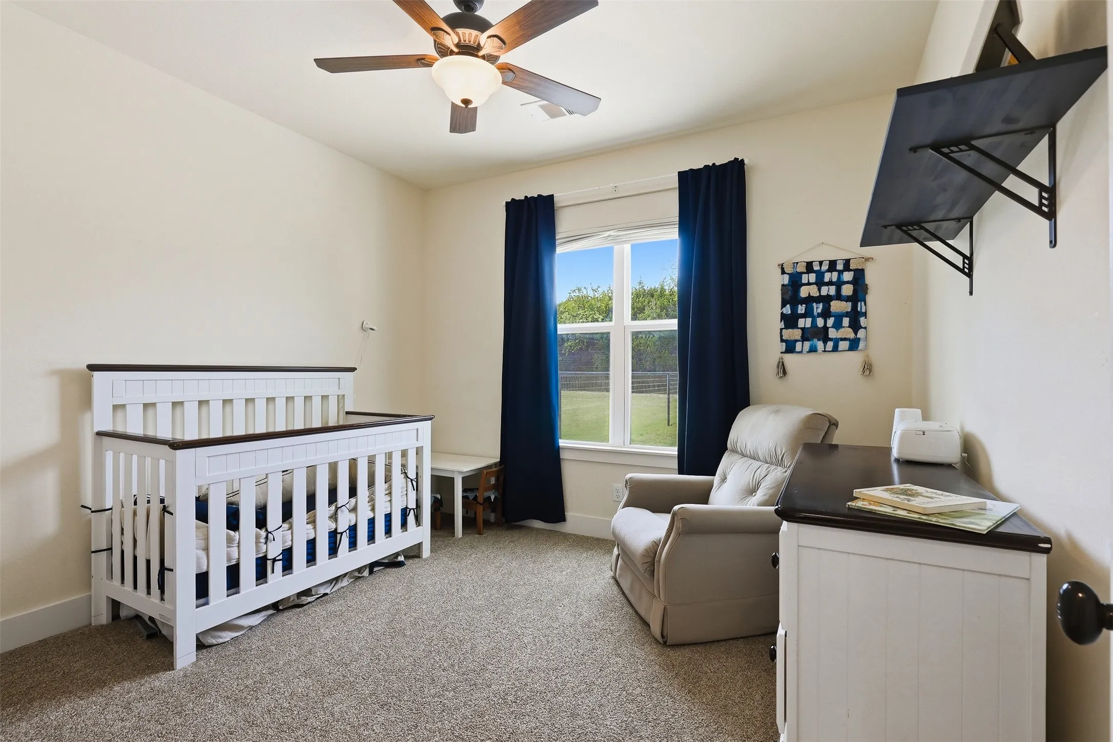 Carpeted room featuring a ceiling fan with integrated lighting, a window with dark blue drapes, and a wall-mounted shelving unit