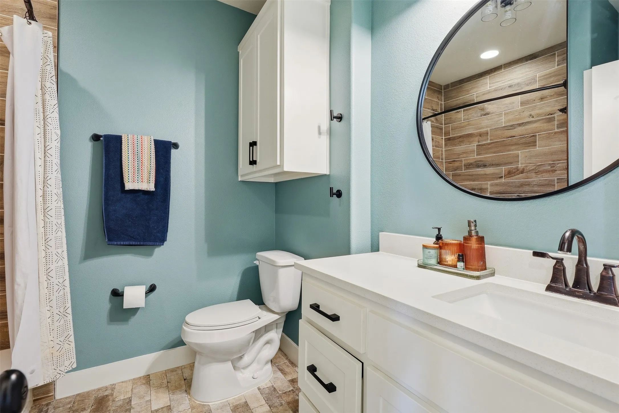 Bathroom featuring a white vanity with integrated sink, a large round mirror with a black frame, light blue walls, and wood-look tile flooring