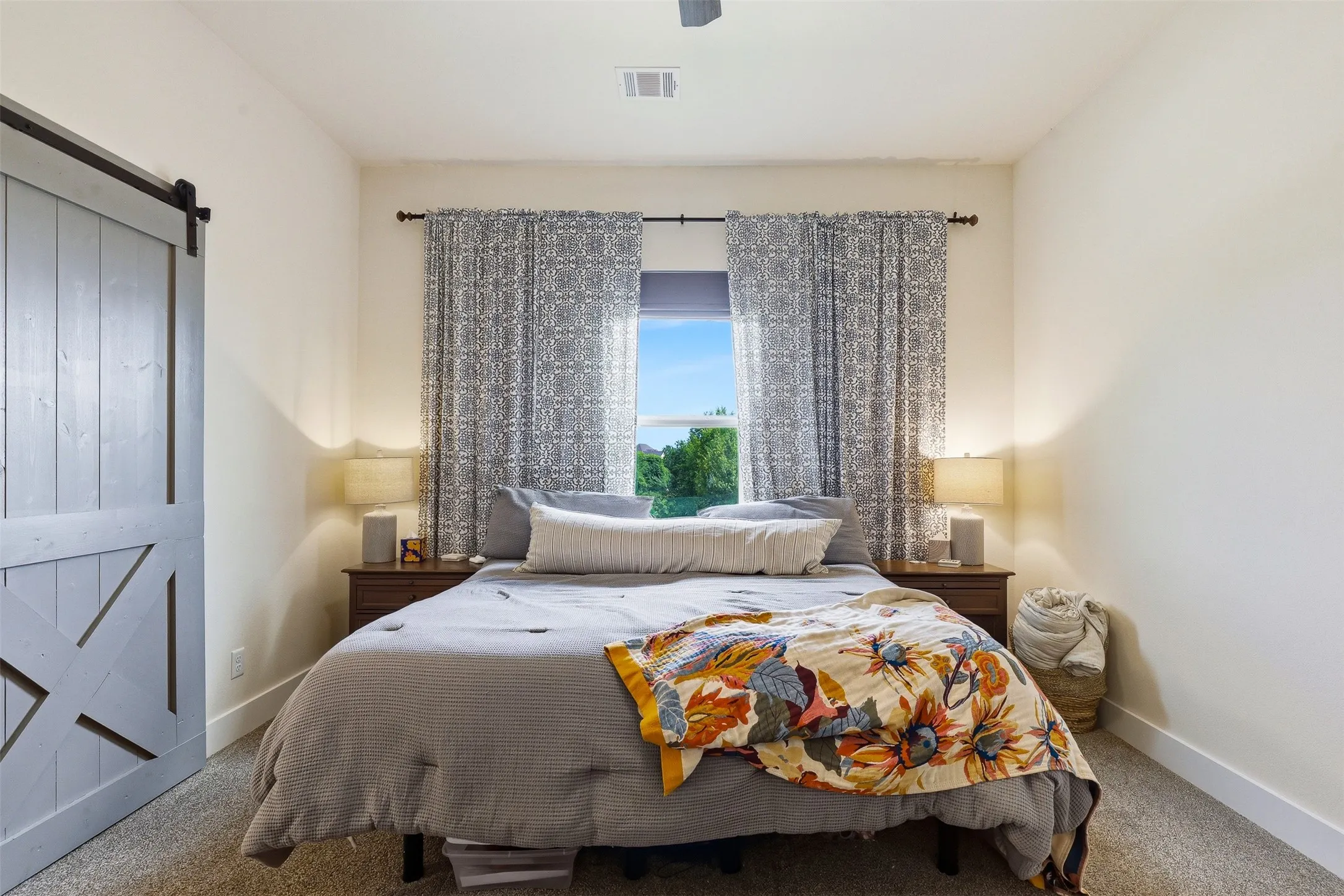 Carpeted room featuring a window with patterned drapes, a ceiling fan, and a light-toned barn door