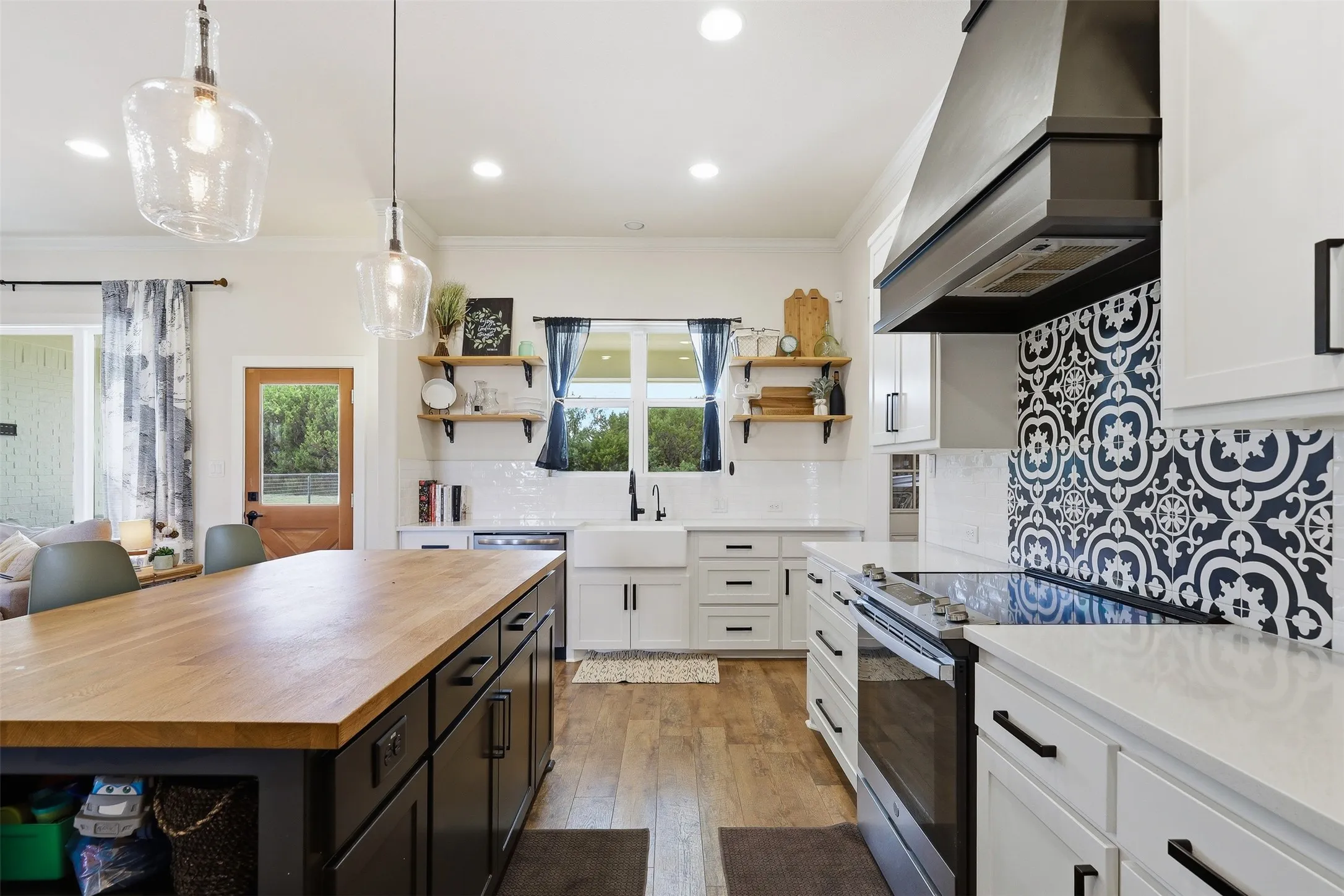 Expansive kitchen with a wood-finish island countertop, white cabinetry with black hardware, patterned tile backsplash, apron-front sink, and custom range hood