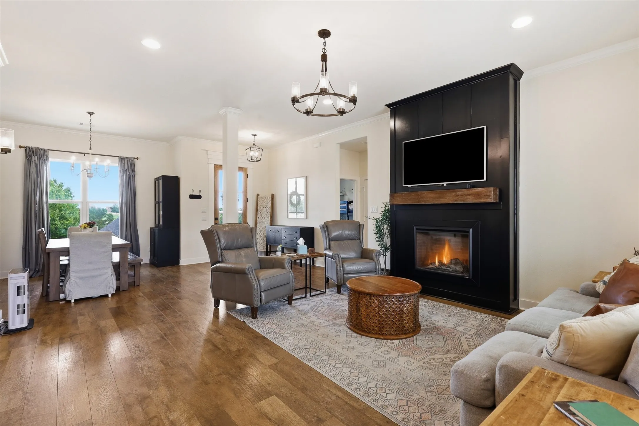 Open-concept living space featuring wood-finish flooring, a contemporary fireplace with a dark paneled surround and wood mantel, and an adjacent dining area with a chandelier