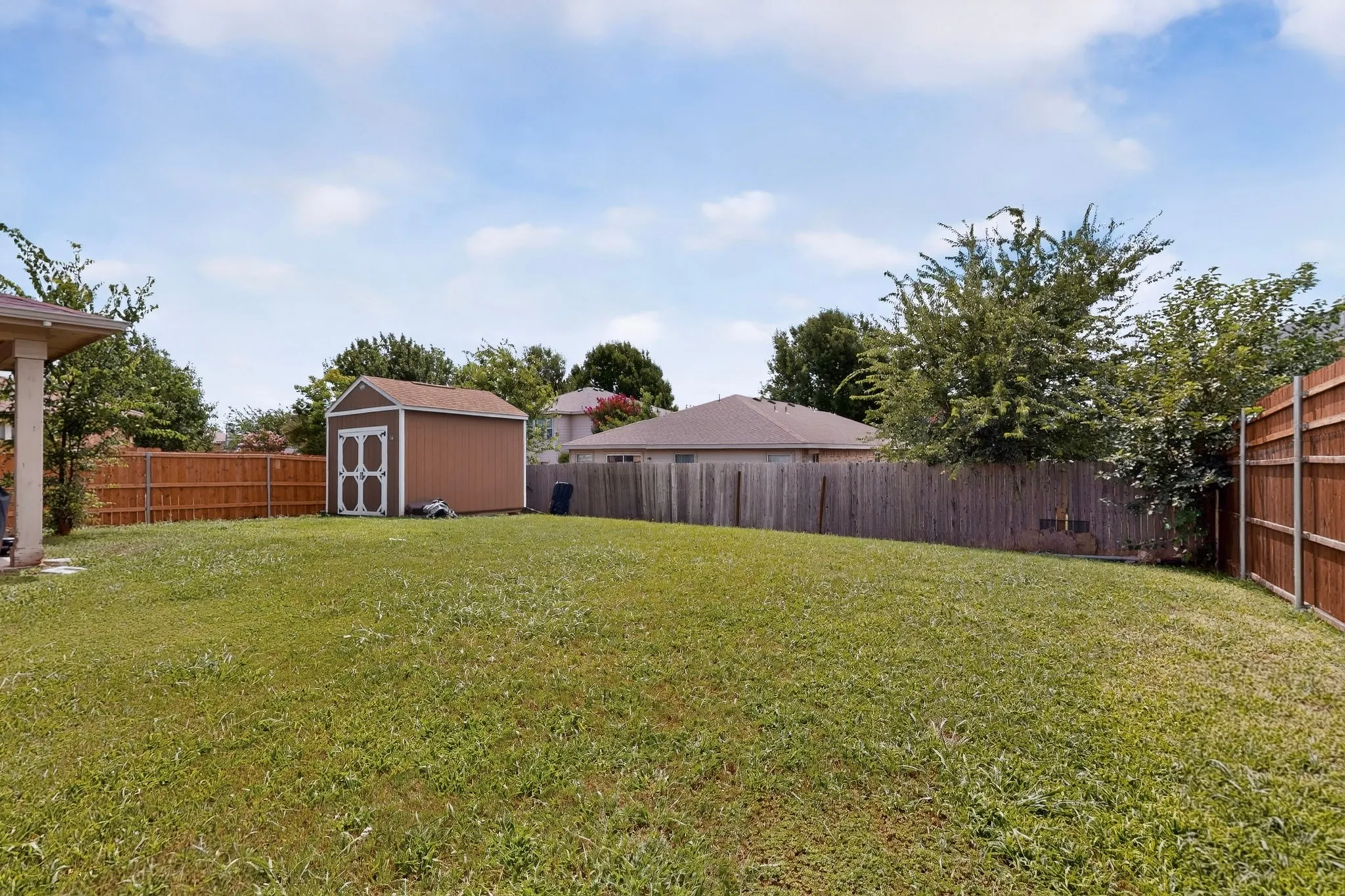 Expansive grassy yard featuring a brown wood storage shed with white trim, full wood privacy fencing, and mature perimeter trees