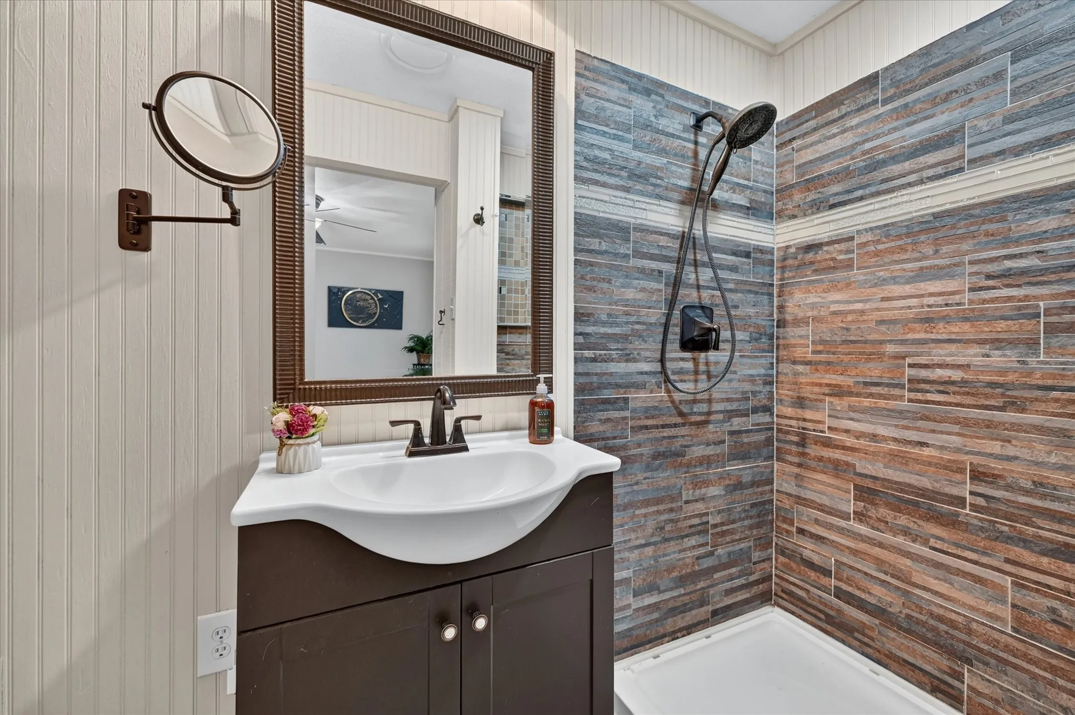 Bathroom featuring beadboard walls, a dark wood-finish vanity with an integrated sink, a framed wall mirror, a wall-mounted magnifying mirror, and a shower with multi-toned tile walls and a matte black shower fixture