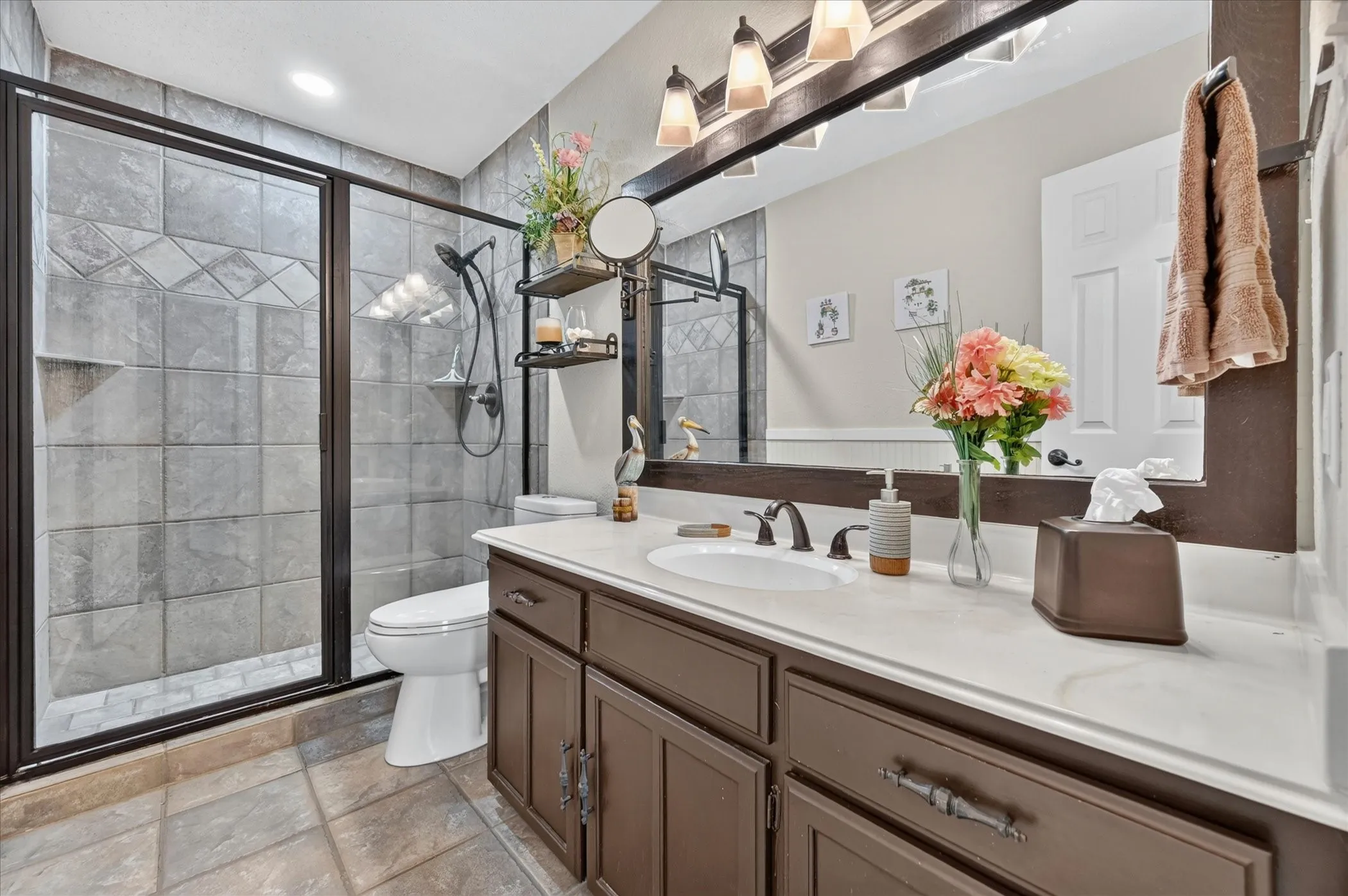 Bathroom featuring a glass-enclosed shower with tile surround, a single vanity with a white countertop and integrated sink, dark wood-finish cabinetry with decorative hardware, and ceramic tile flooring