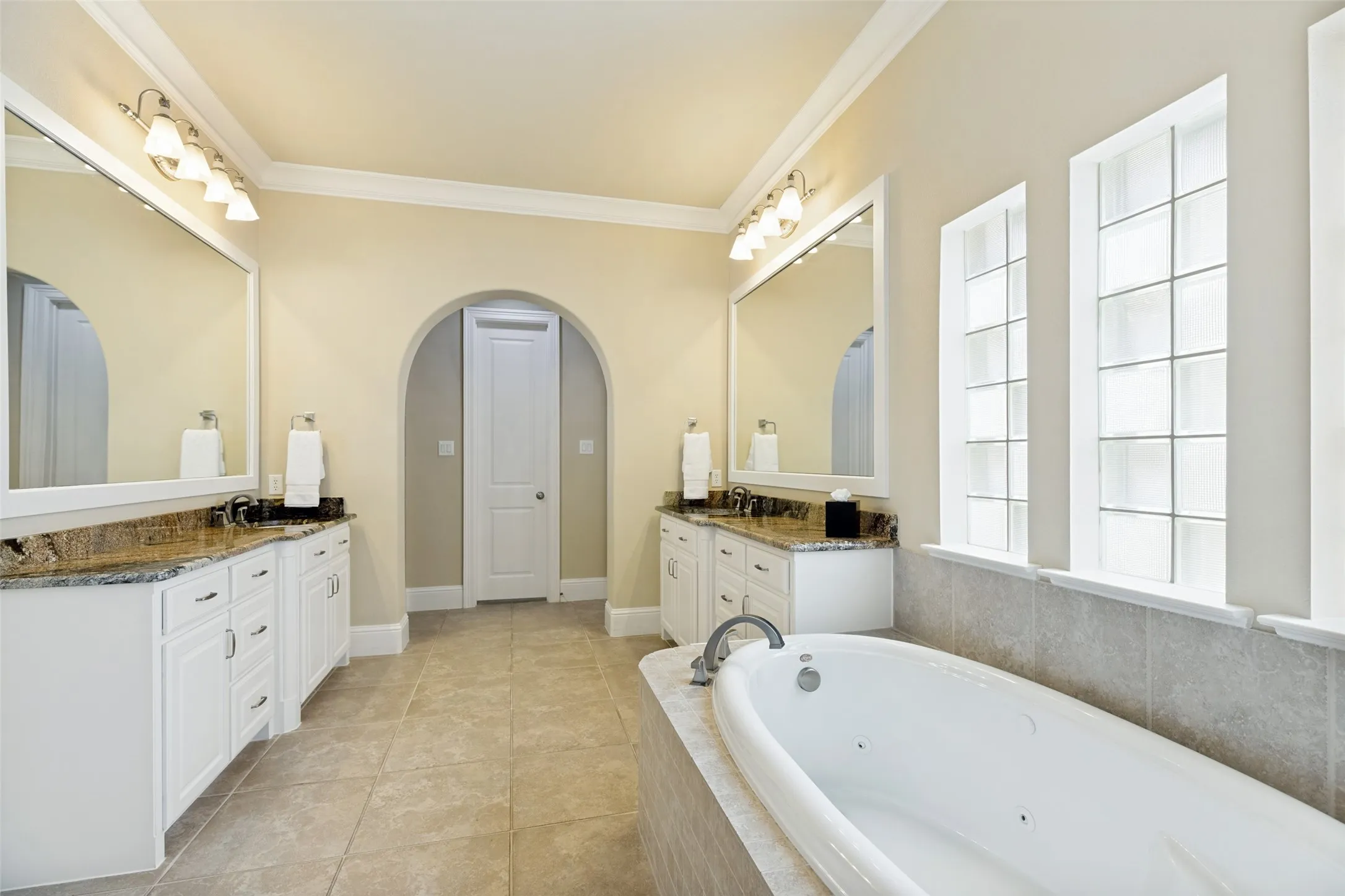 The elegant primary bath provides a pair of wide white vanity cabinets topped by contrasting granite countertops with undermount sinks.  White framed mirrors hang above the vanities. Pretty lights topping the mirrors and a trio of windows with obscure glass all illuminate the cheerful bath.  A jetted drop-in tub is below the windows.