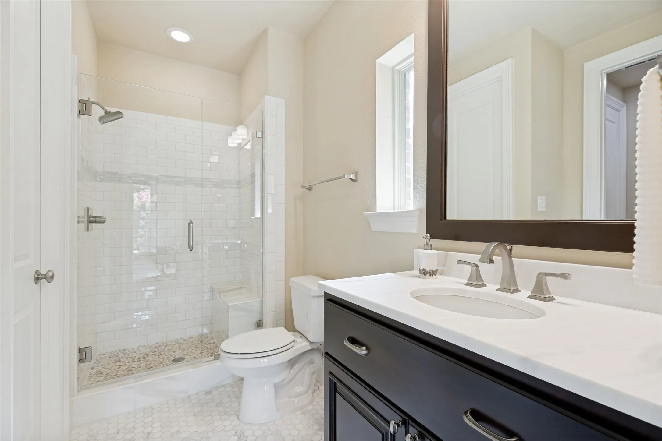 This attractive bath opens to the office and to the hallway for guests.  The dark wood vanity is topped by a cultured marble countertop with an undermount sink.  A framed mirror is above.  Offset subway tile surrounds the shower and it has a frameless glass enclosure.  A smooth pebble mosaic is on the floor of the shower, and a handy bench is included.  Small octagonal tiles are on the floor and a window and fixture above the mirror provide light. Brushed nickel fixtures and hardware provide the finishing touches.