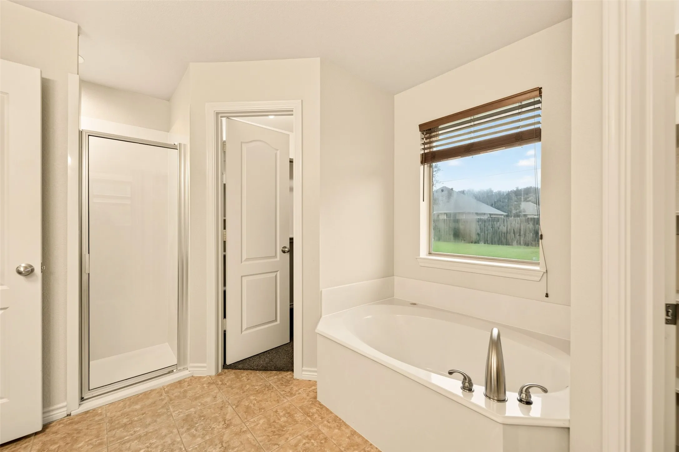 Corner soaking tub with brushed nickel fixtures, a private window with wood-finish blinds, and a tiled floor, and walk-in closet #1.