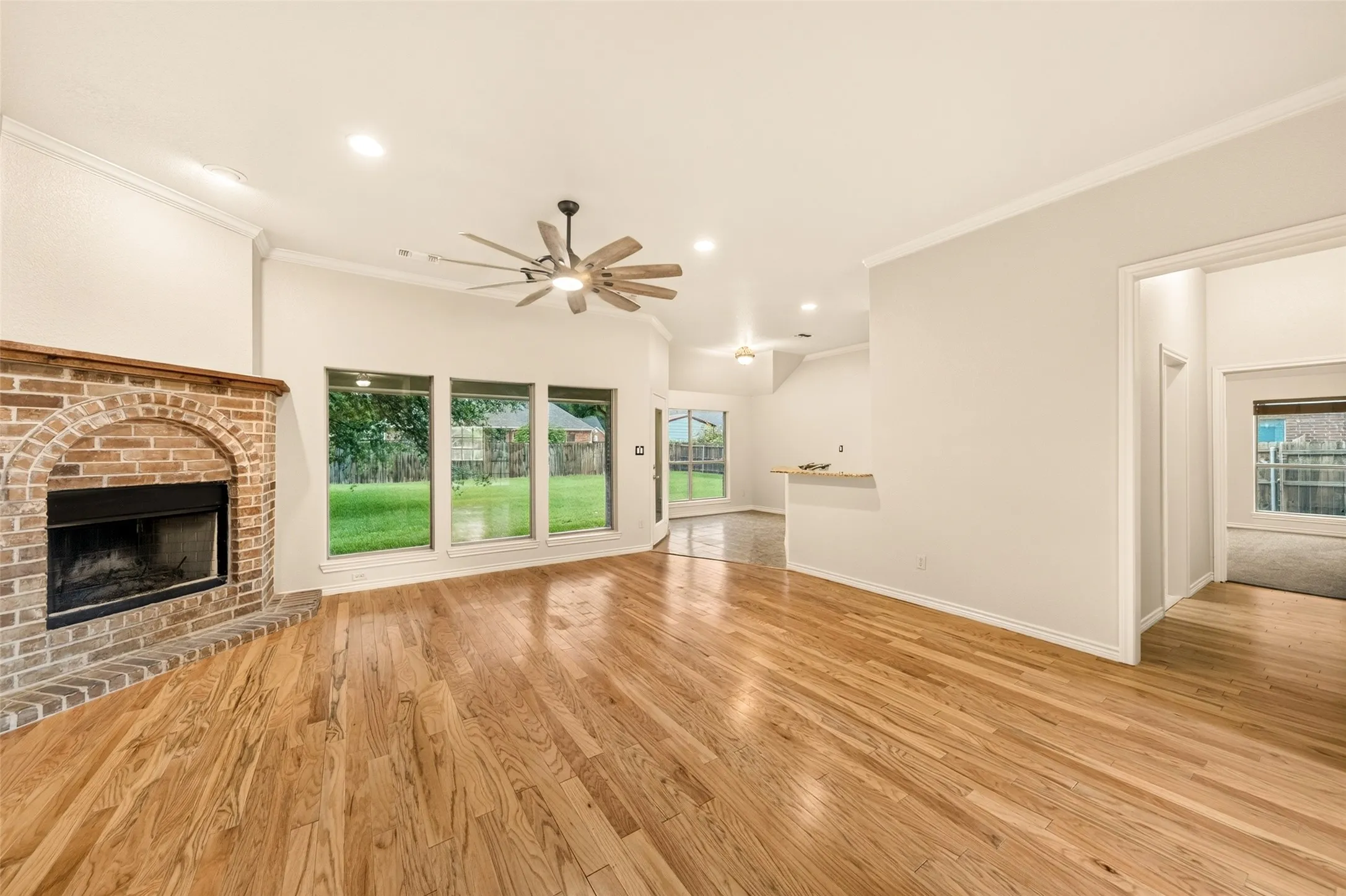 Spacious living area featuring wood-finish flooring, a brick fireplace with a wood mantel, and large windows overlooking the​​‌​​​​‌​​‌‌​‌‌​​​‌‌​‌​‌​‌​​​‌​​ backyard