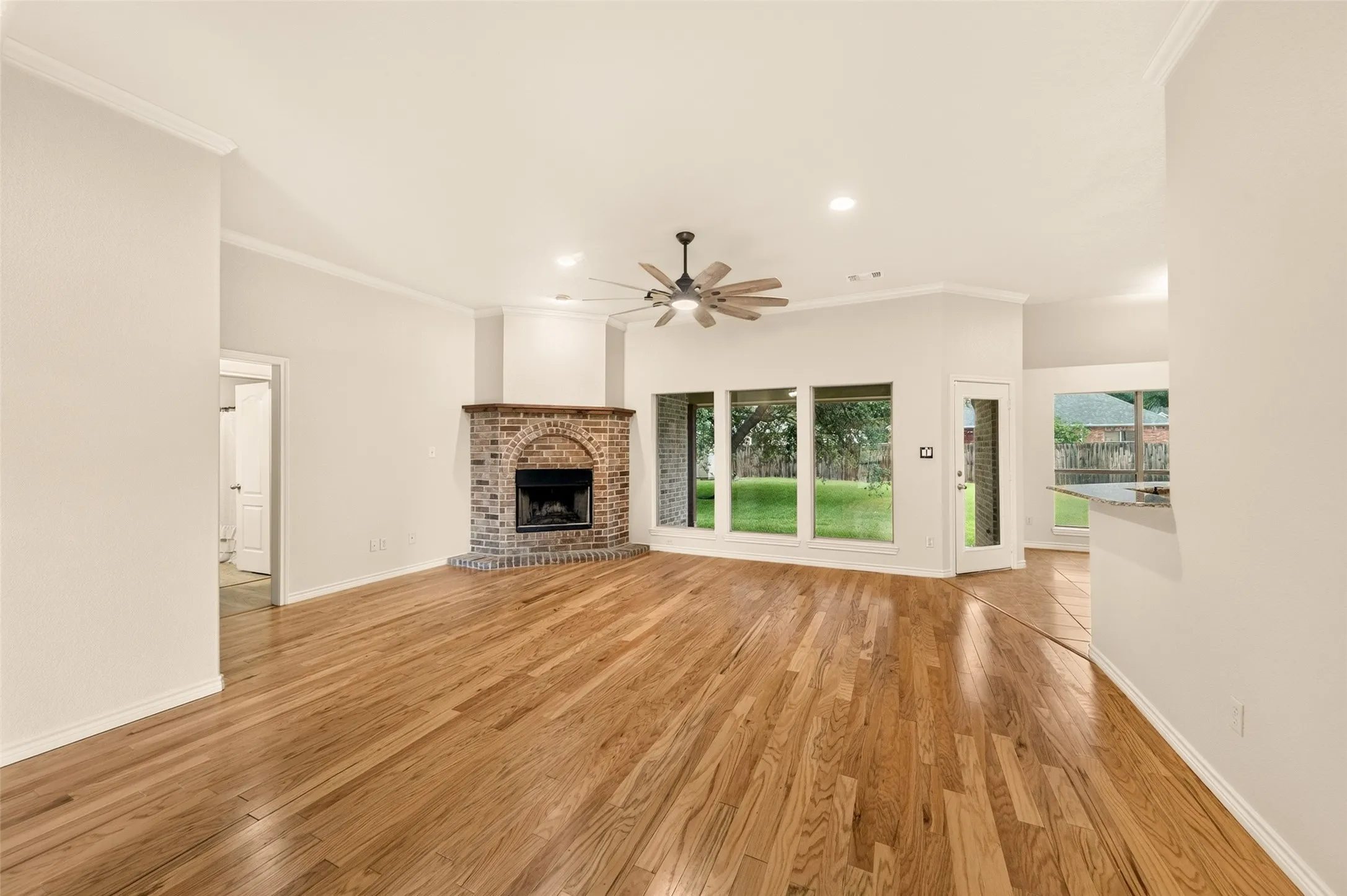 Spacious living area featuring wood-finish flooring, a brick fireplace with an arched opening, multiple large windows, a ceiling fan with integrated lighting, and crown molding