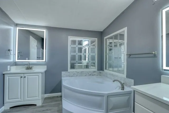 Corner soaking tub with a tiled surround, two framed obscure glass windows, and illuminated vanity mirrors