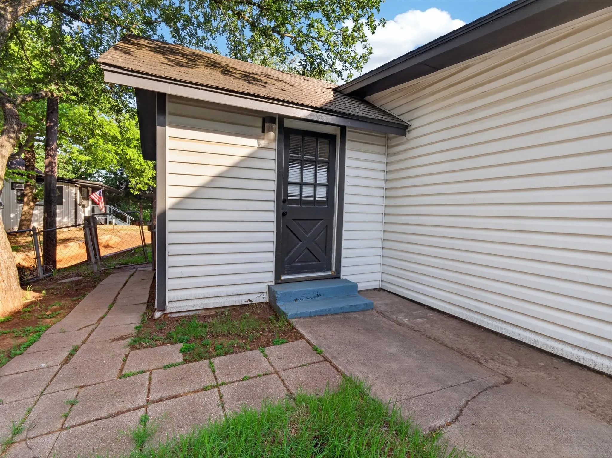 White horizontal siding exterior with a dark-paneled door, concrete walkway, and paved patio area