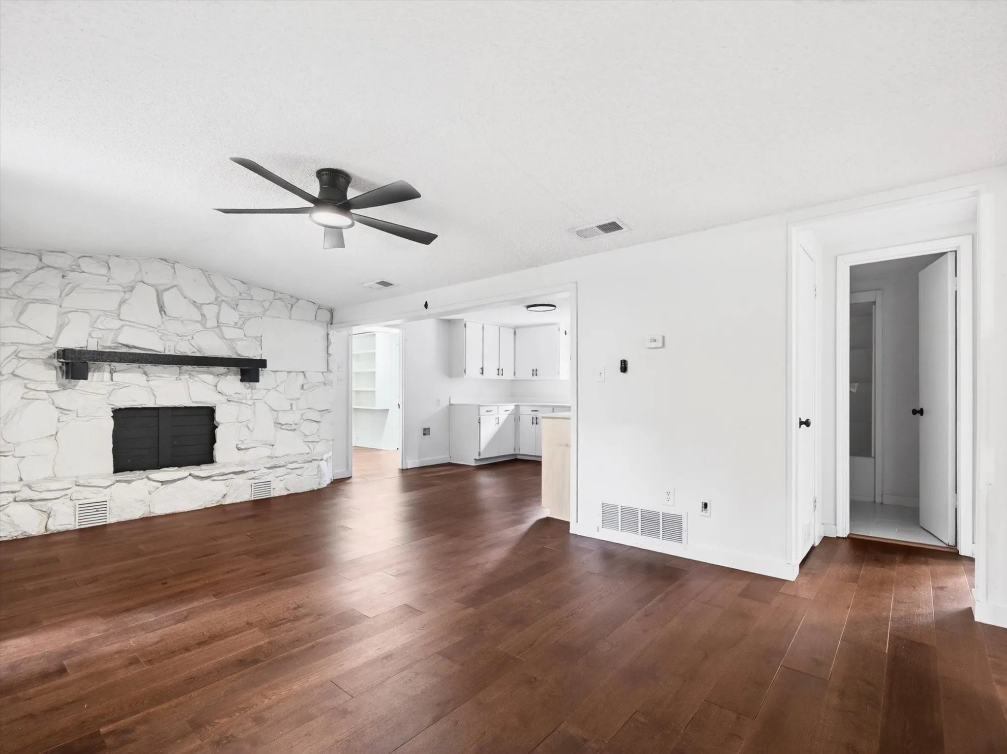 Spacious living area featuring a stone-clad fireplace with a dark wood mantel, wood-finish flooring, a contemporary ceiling fan, and white walls