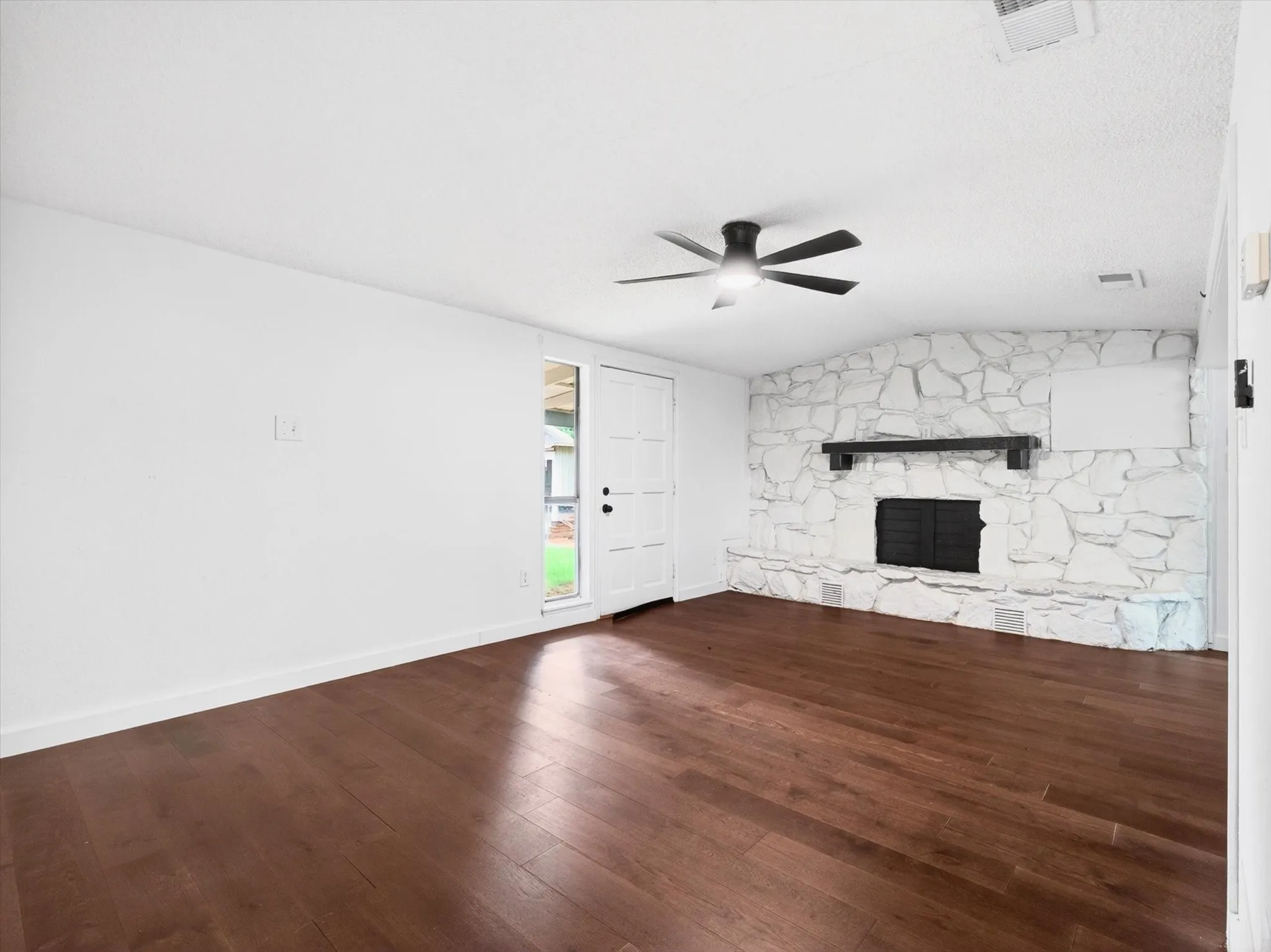 Living space featuring rich wood-finish flooring, a white painted stone fireplace with a dark mantel, and a modern ceiling fan