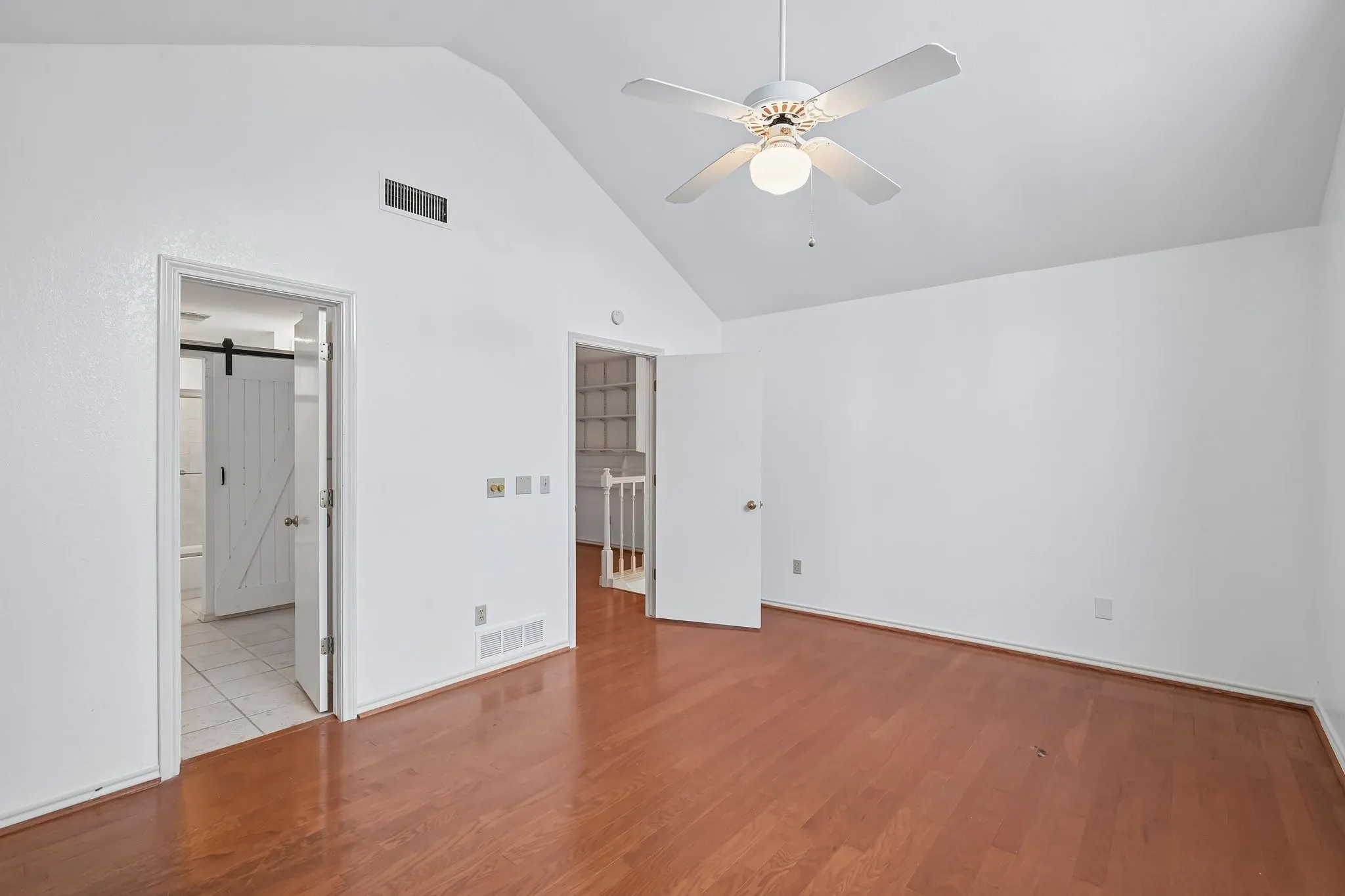 Spacious room featuring vaulted ceilings, wood-finish flooring, a ceiling fan with light fixture, and a barn door entry to a tiled area