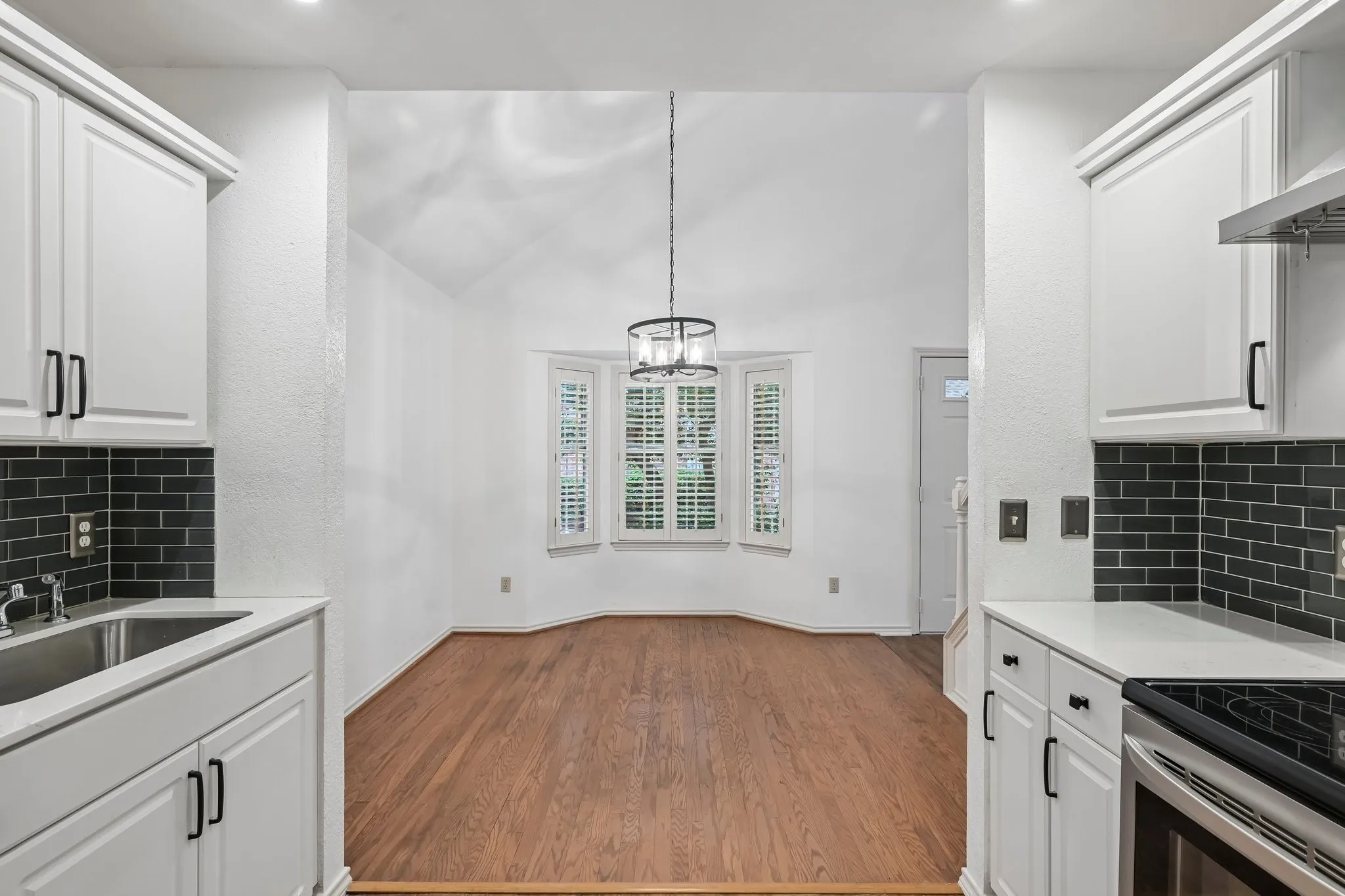 Kitchenette featuring white cabinetry, dark subway tile backsplash, and a stainless steel sink, opening into a dining area with wood-finish flooring, a vaulted ceiling, and a bay window with plantation shutters
