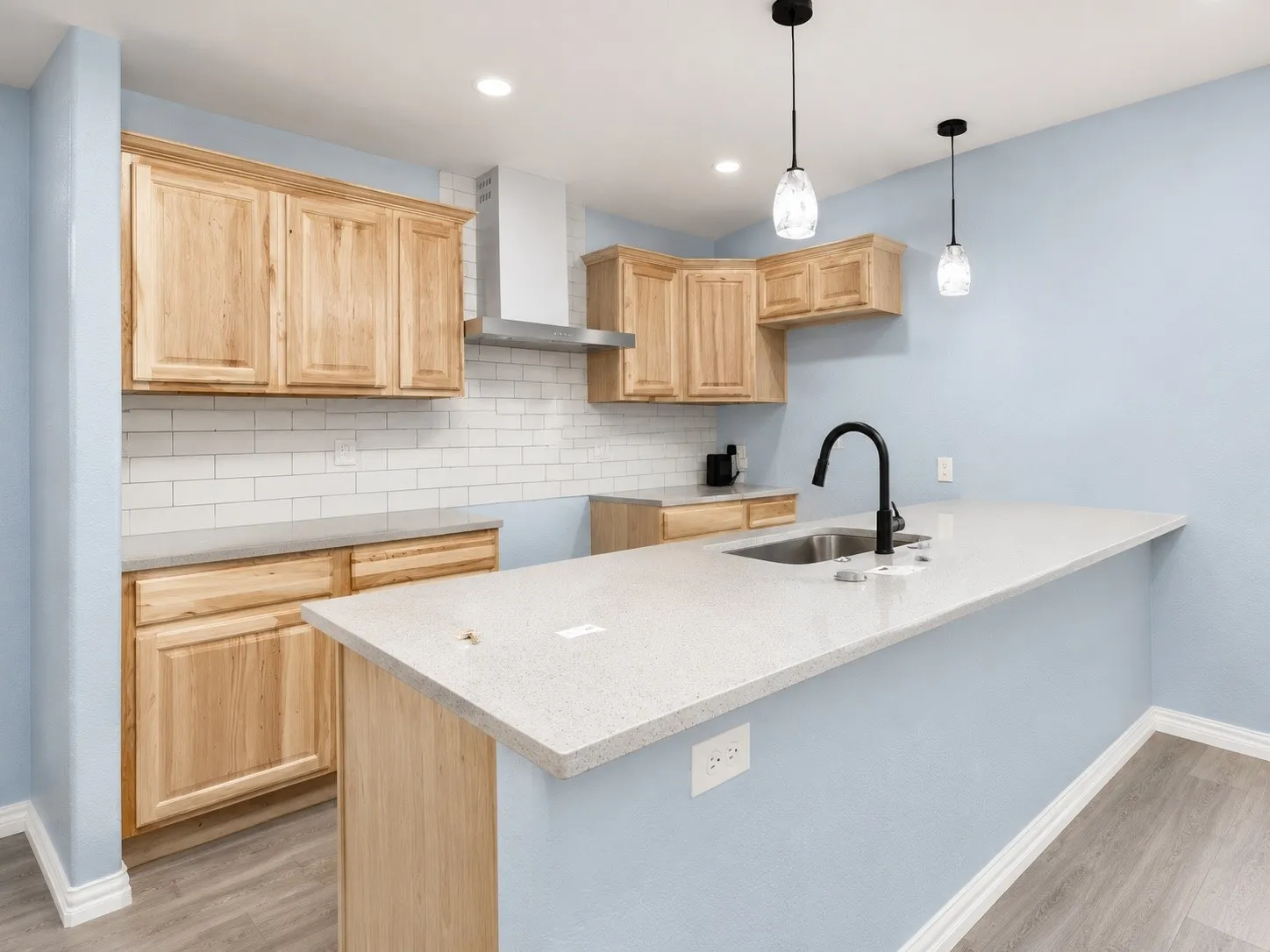 Kitchen featuring light wood cabinetry, white subway tile backsplash, light-toned countertops, and a stainless steel sink with a matte black faucet