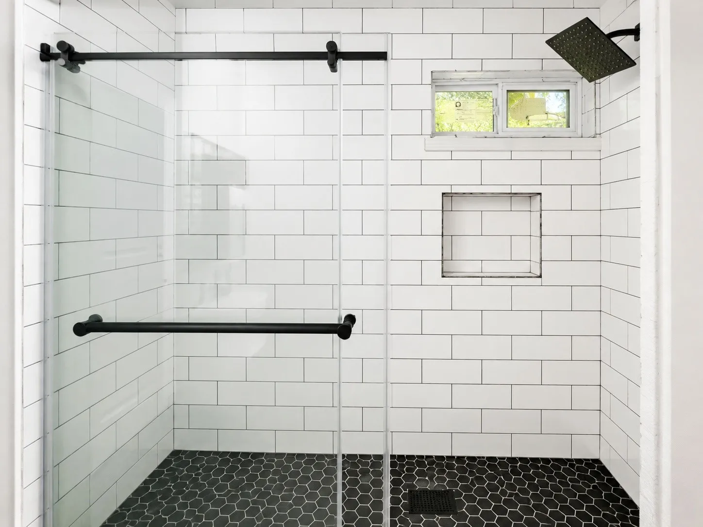 Modern shower enclosure featuring clear glass panels with matte black hardware, white subway tile walls, a hexagonal mosaic tile floor, an integrated shower niche, and a fixed rainfall showerhead