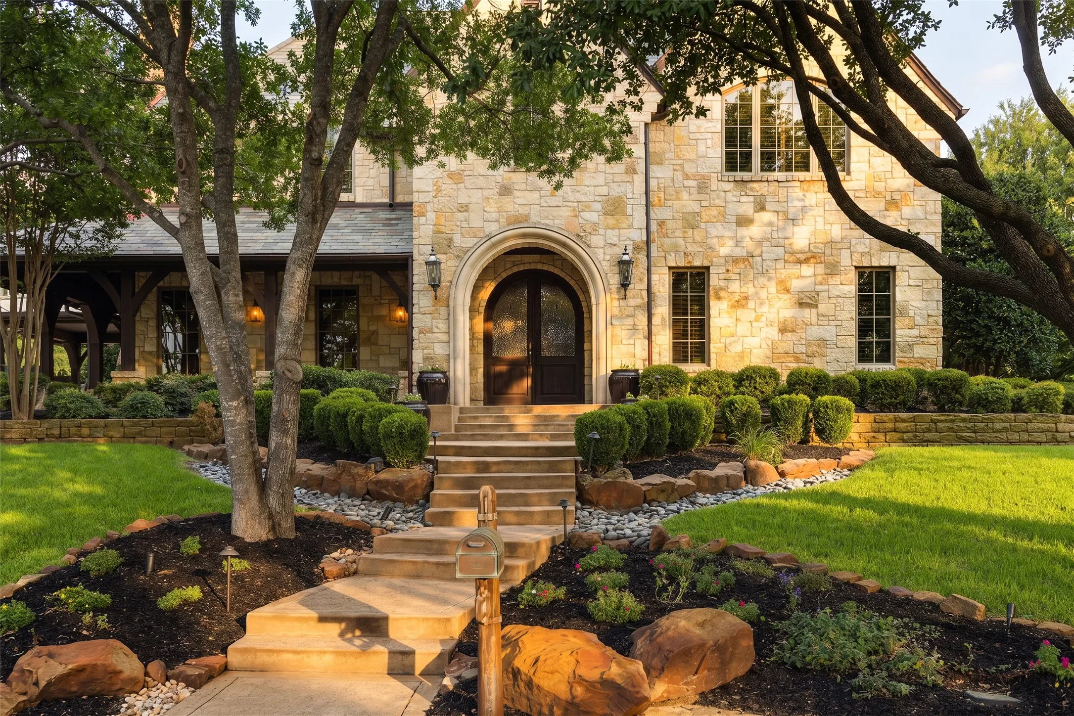 Stone facade residence featuring an arched entryway with double doors, symmetrical windows, a terraced landscape with established plantings, and a stone retaining wall