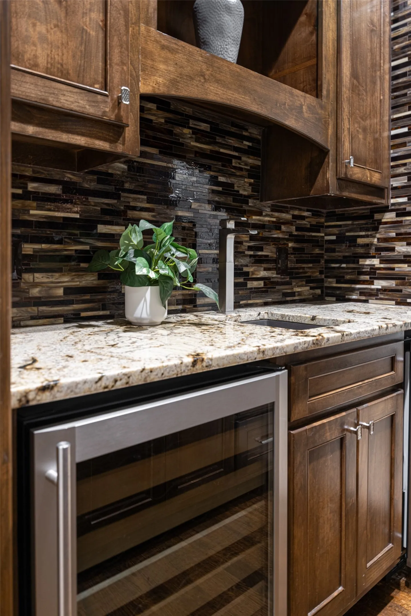 Built-in beverage cooler beneath a granite-style countertop, complemented by rich wood cabinetry and a mosaic tile backsplash