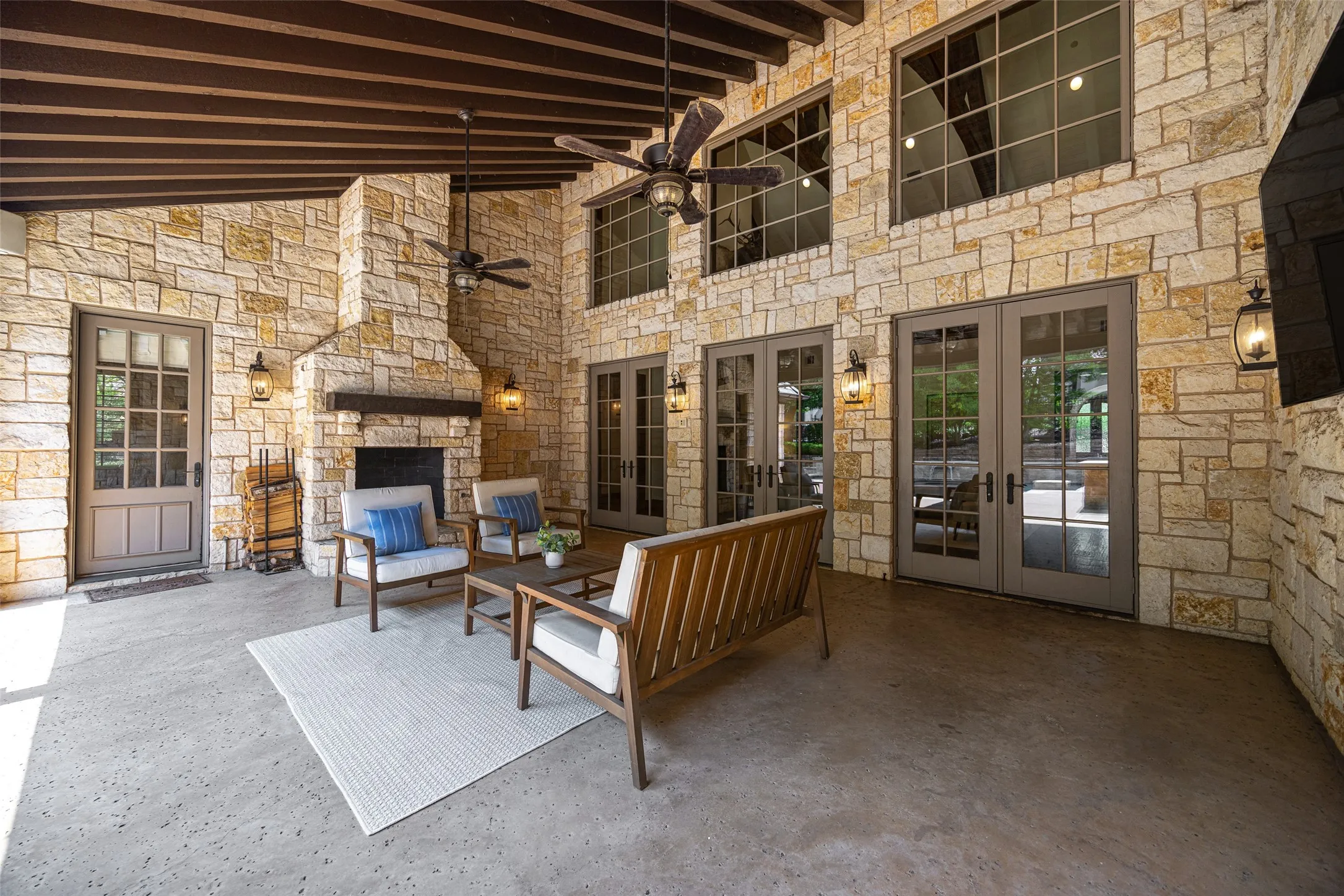 Spacious outdoor living area with stone walls, exposed beam ceiling, a built-in fireplace with a wood mantle, and multiple French doors