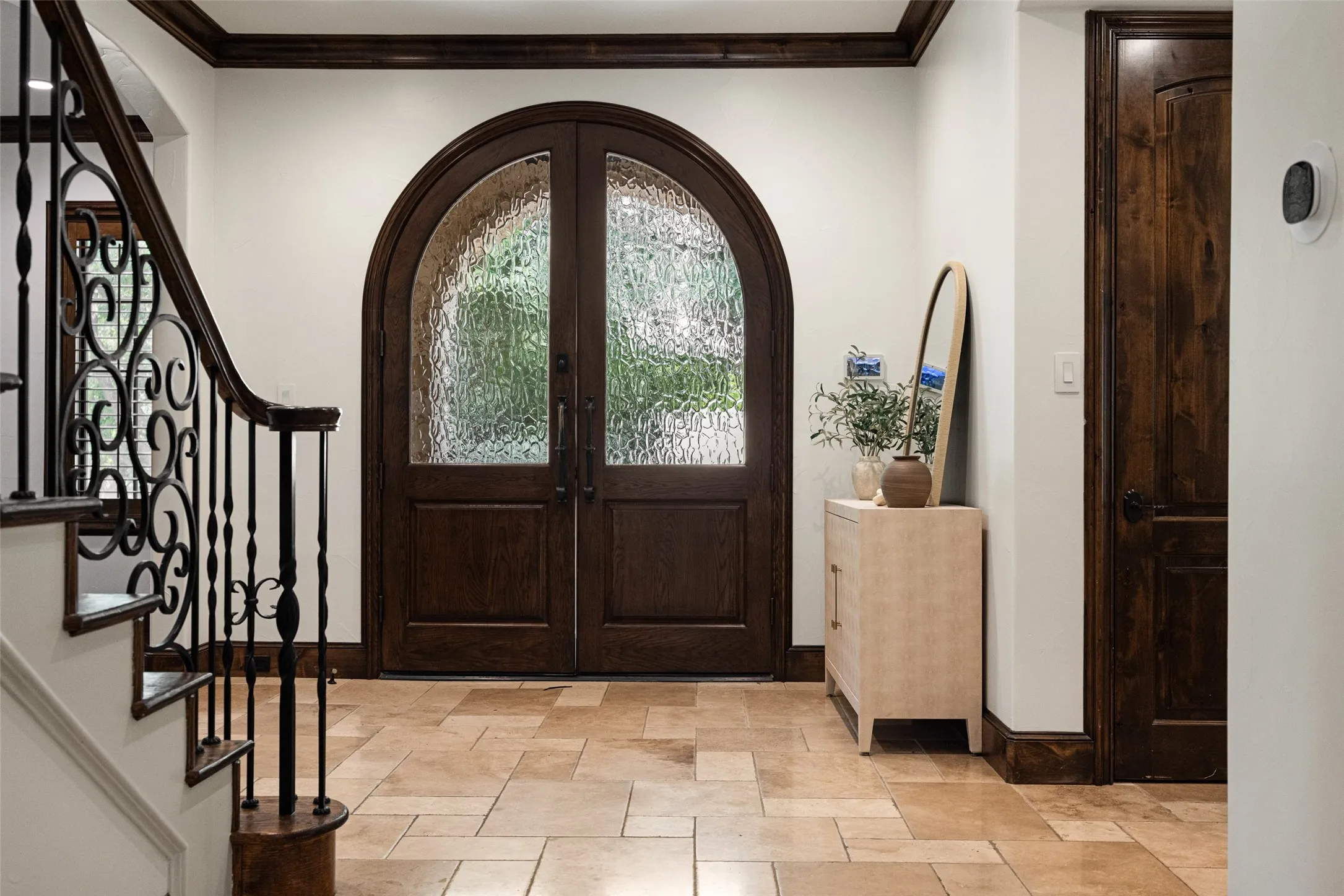 Grand arched double entry doors with textured glass panels, dark wood trim, tile flooring, and a curved staircase with wrought iron balusters