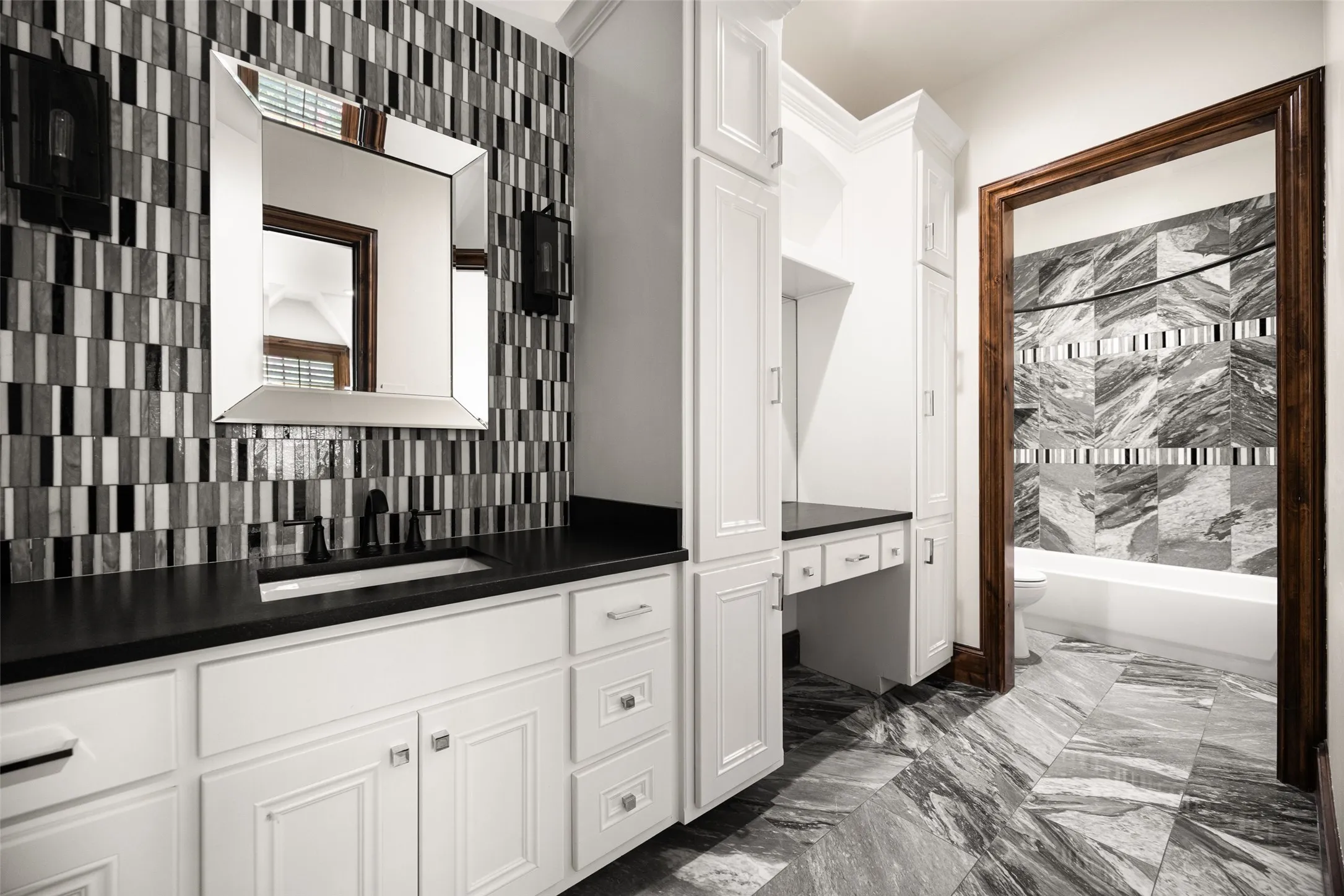 Vanity area featuring white cabinetry, a dark countertop, an integrated rectangular sink, a decorative tiled backsplash, and an ornate framed mirror