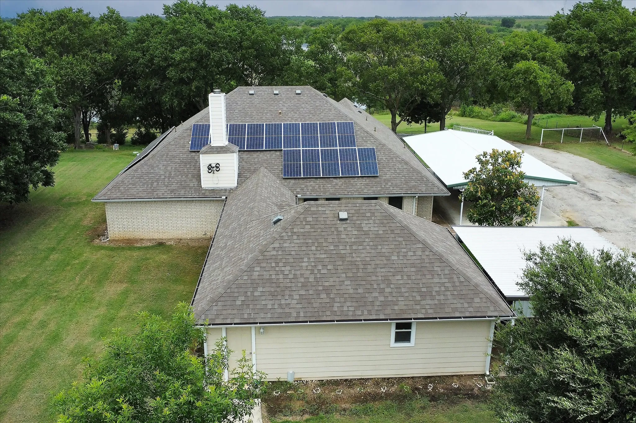 Shingle roof with integrated solar panels, brick and siding exterior, white chimney, and two white metal outbuildings