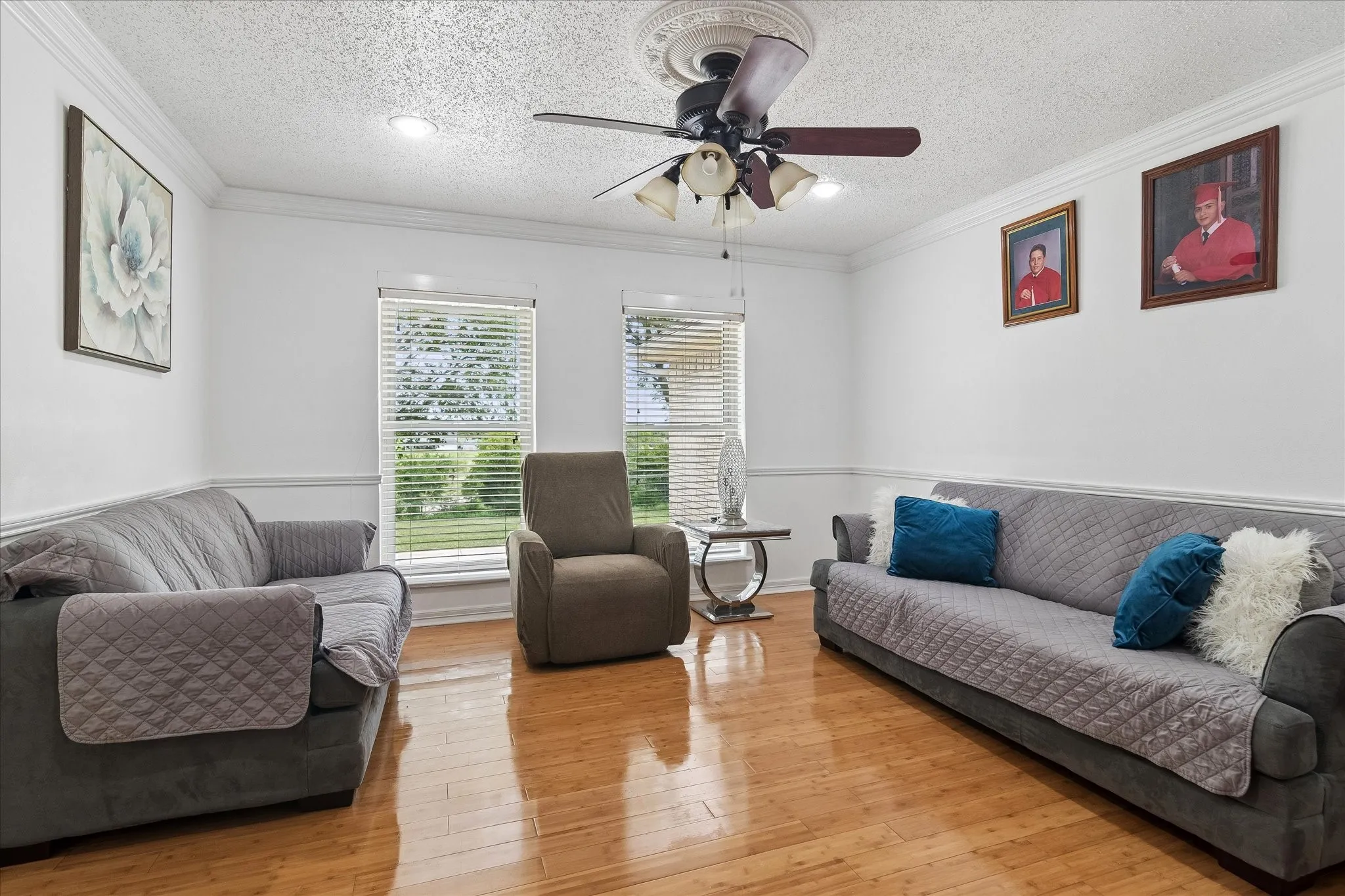Living area featuring wood-finish flooring, two large windows with horizontal blinds, a ceiling fan with light kit, recessed lighting, and crown molding