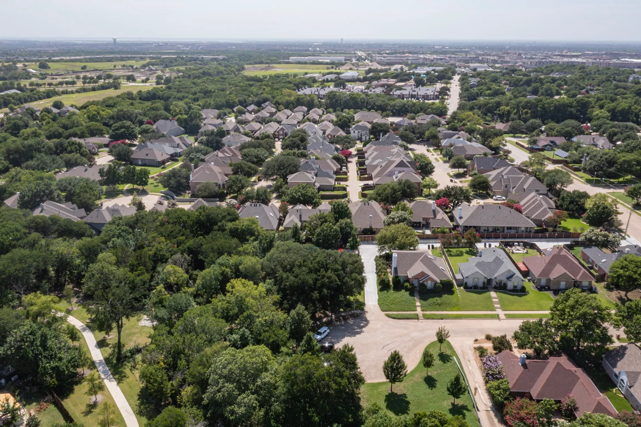Aerial perspective of a residential community featuring numerous single-family homes with pitched roofs, surrounded by mature trees