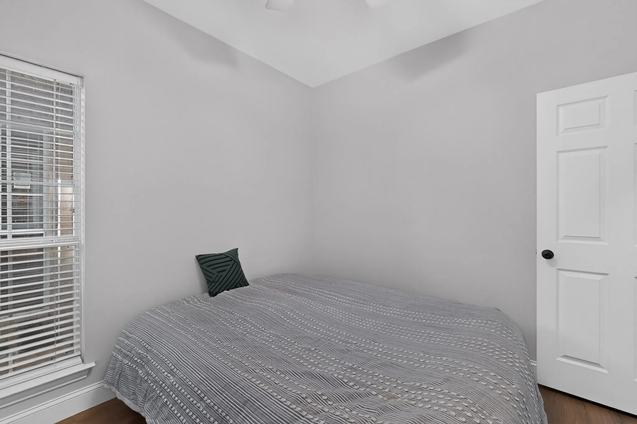 Neutral-toned room featuring a single window with blinds, a white paneled door with a black knob, and wood-finish flooring
