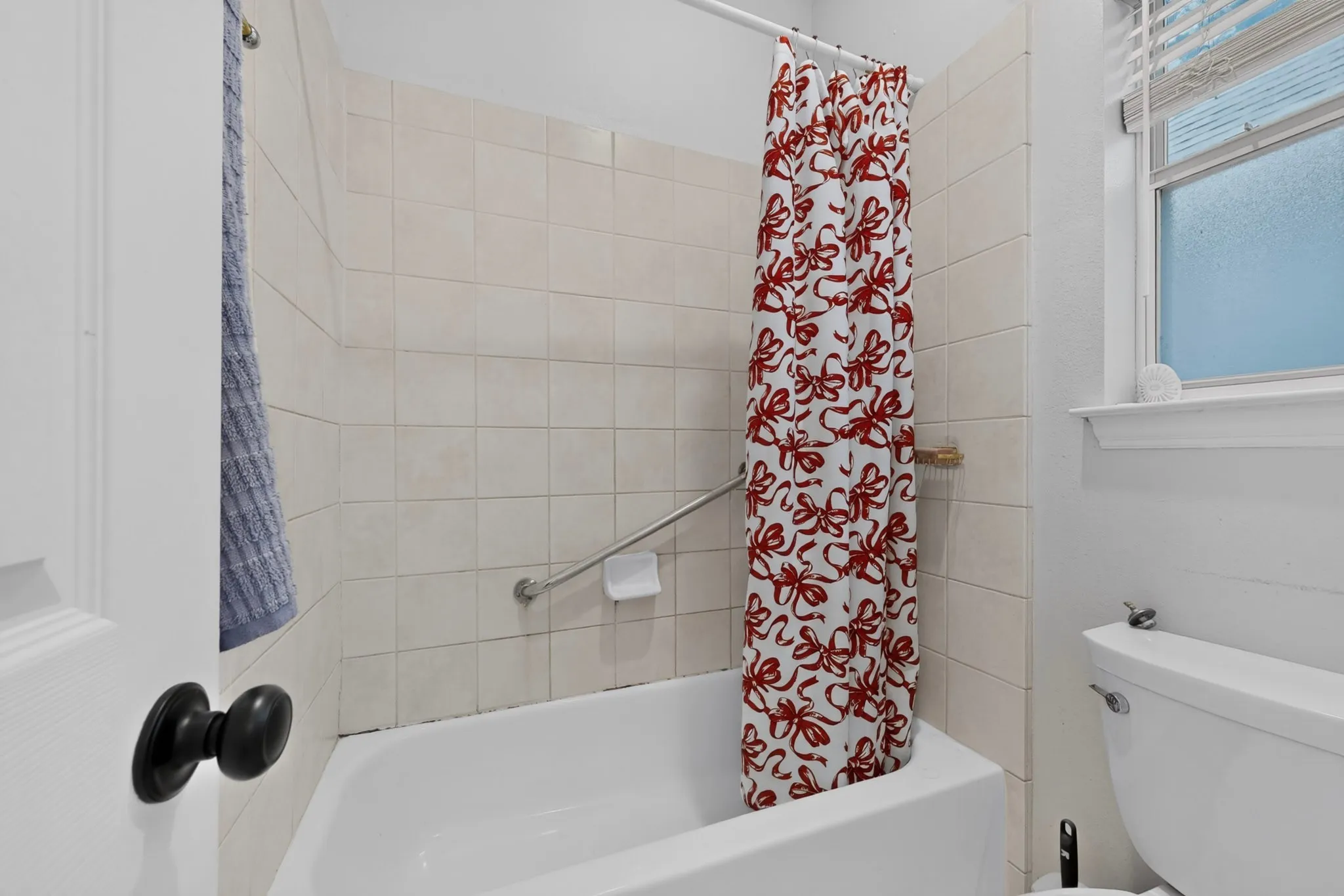 Bathroom featuring a white bathtub, ceramic tile surround, built-in grab bar, and a window with privacy blinds