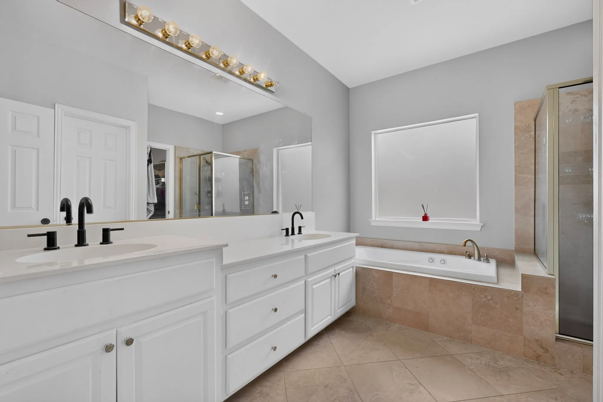 Bathroom featuring a dual vanity with white cabinetry and countertops, matte black plumbing fixtures, a built-in soaking tub with tiled surround, and a glass-enclosed shower