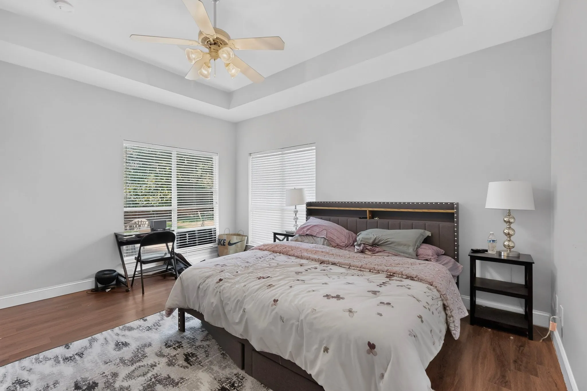 Spacious room featuring a tray ceiling with a ceiling fan, dual windows providing natural light, wood-finish flooring, and crisp white baseboards