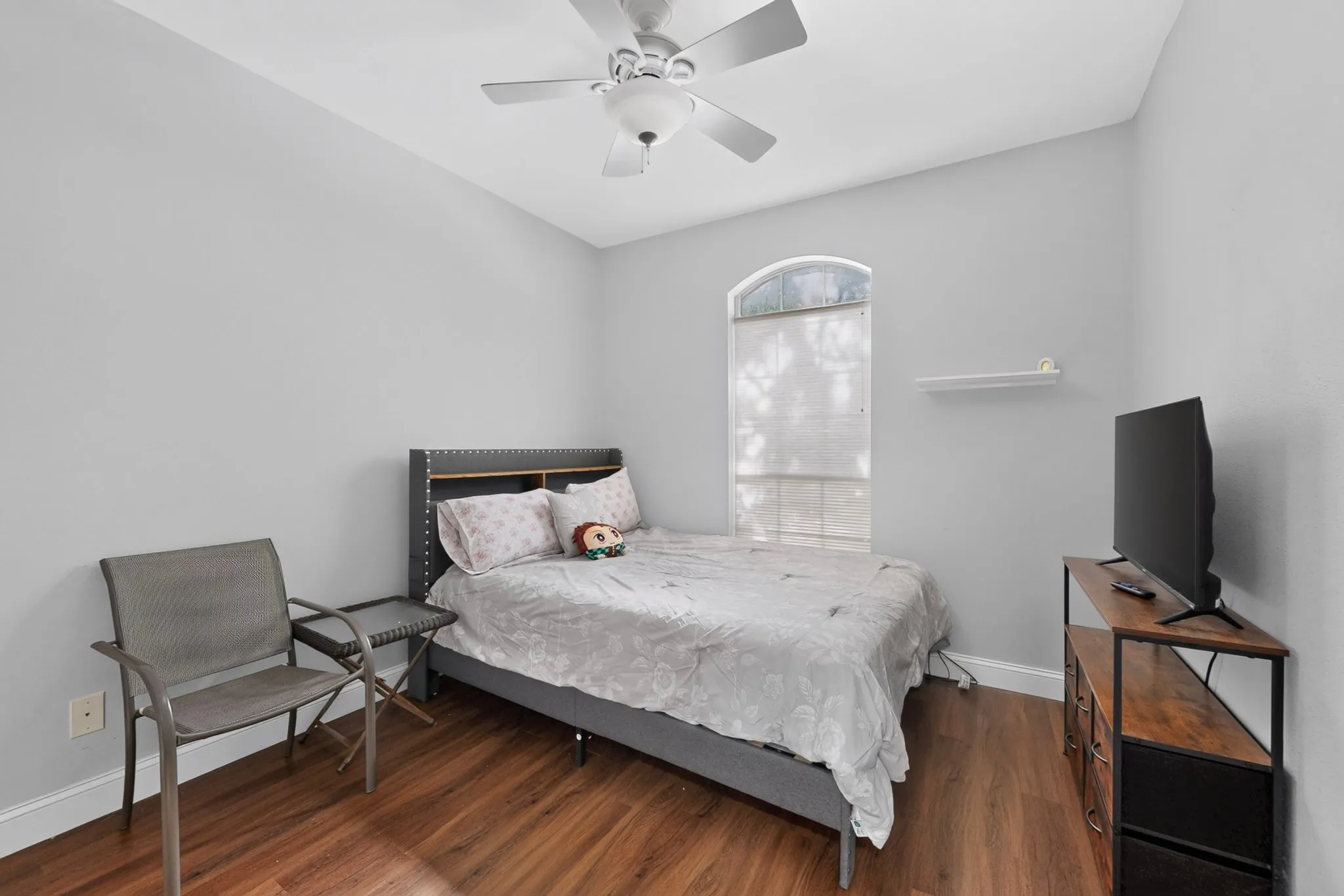Spacious room featuring wood-finish flooring, light gray walls, an arched window, a ceiling fan, and a contemporary floating shelf