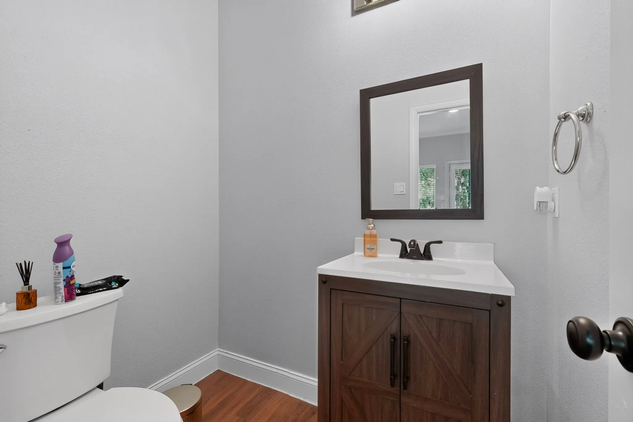 Powder room with wood-finish flooring, a dark wood-finish vanity with a white countertop and integrated sink, a dark framed mirror, and a chrome towel ring