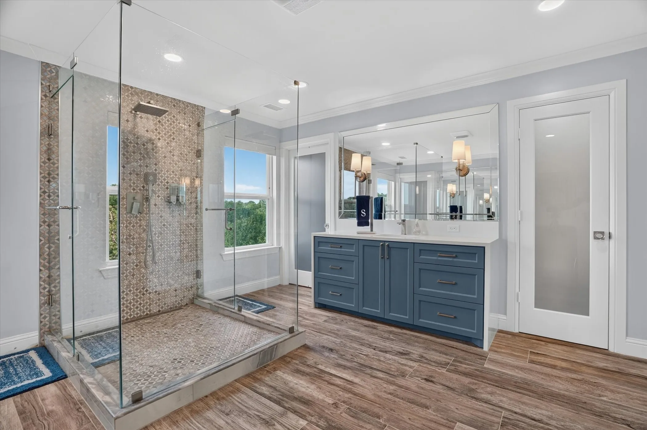 Spacious bathroom featuring a glass-enclosed shower with patterned tile, a double vanity with a quartz countertop and blue cabinetry, wood-finish flooring, and a full-length vanity mirror