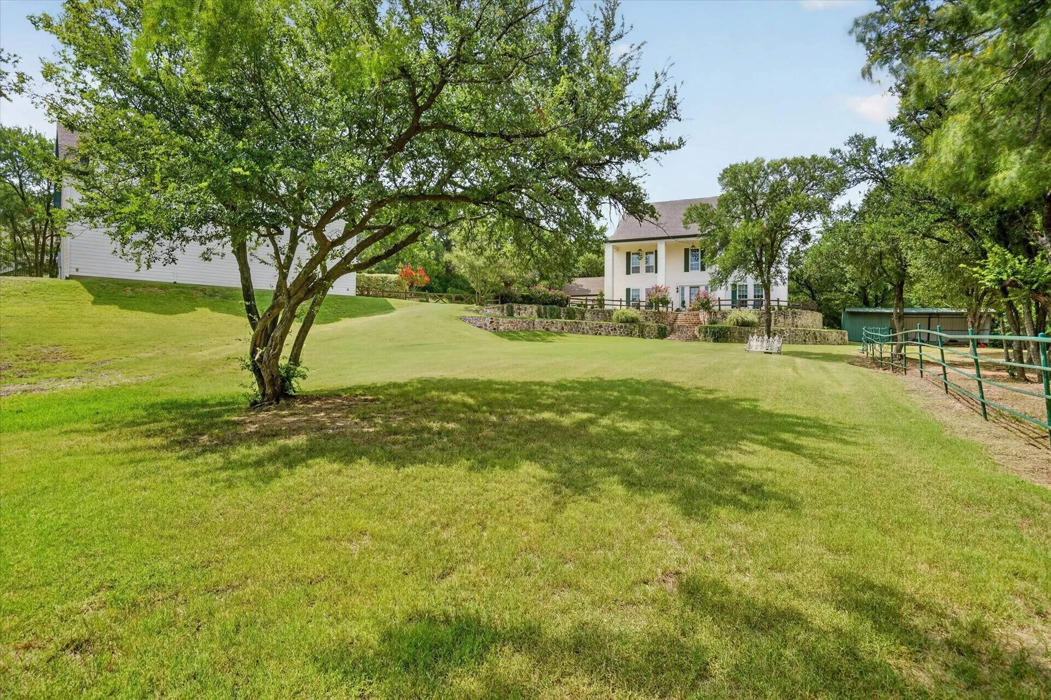 Expansive grassy lawn with mature trees and a stone retaining wall, featuring a traditional-style residence with white siding and dark shutters