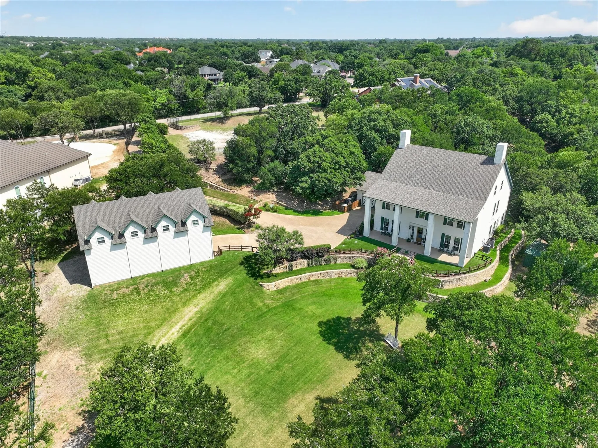 Expansive property featuring a classic two-story residence with a columned front porch, a separate multi-dormered building, tiered landscaping, a gravel driveway, and mature tree coverage