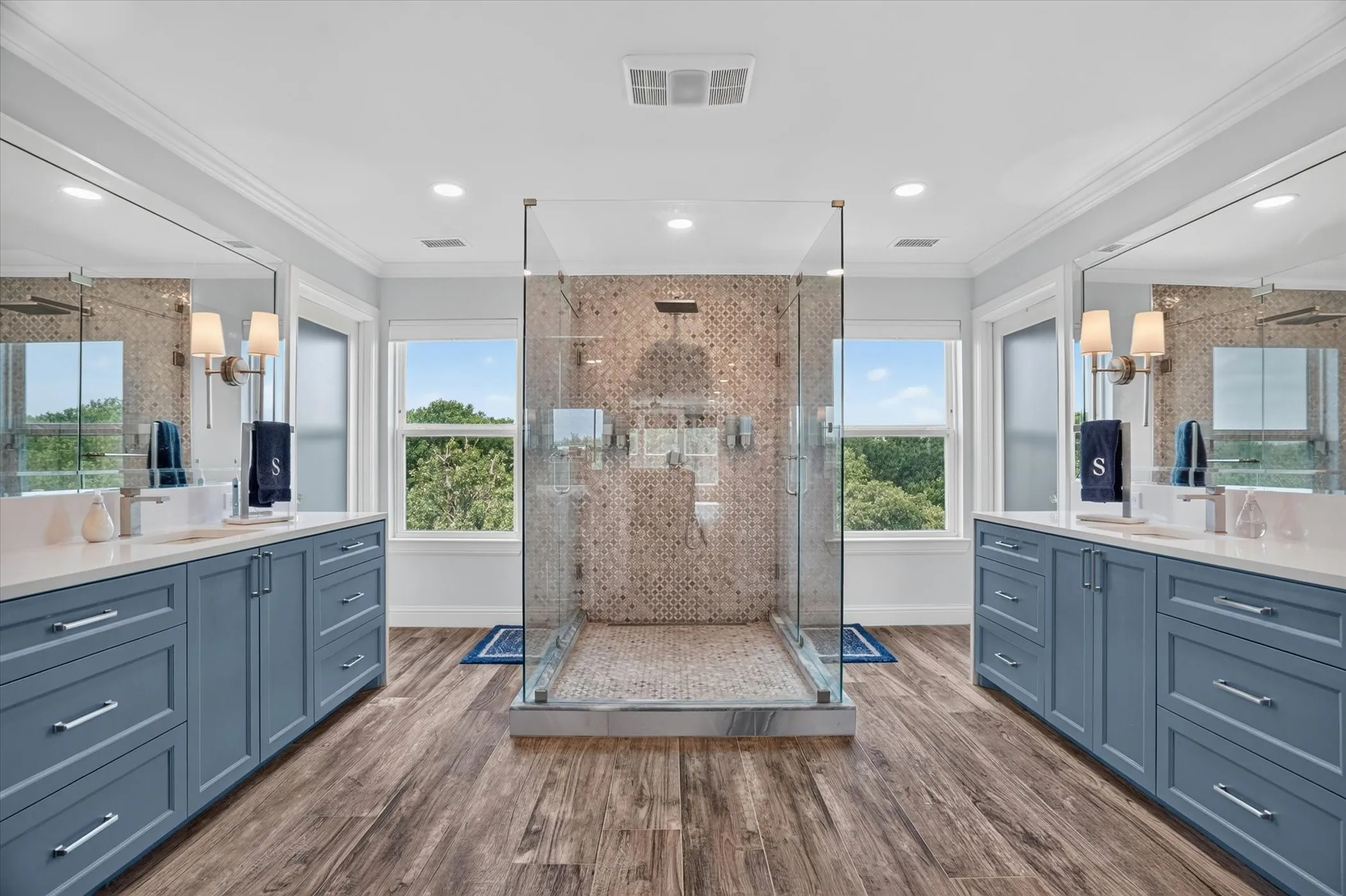 Glass-enclosed shower with mosaic tile surround, flanked by dual vanities featuring blue cabinetry, white countertops, and integrated sinks
