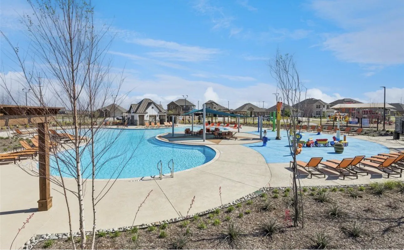 Expansive community pool area featuring a large swimming pool, a dedicated splash pad with water features, a shaded lounging pavilion, and a separate building with a covered patio