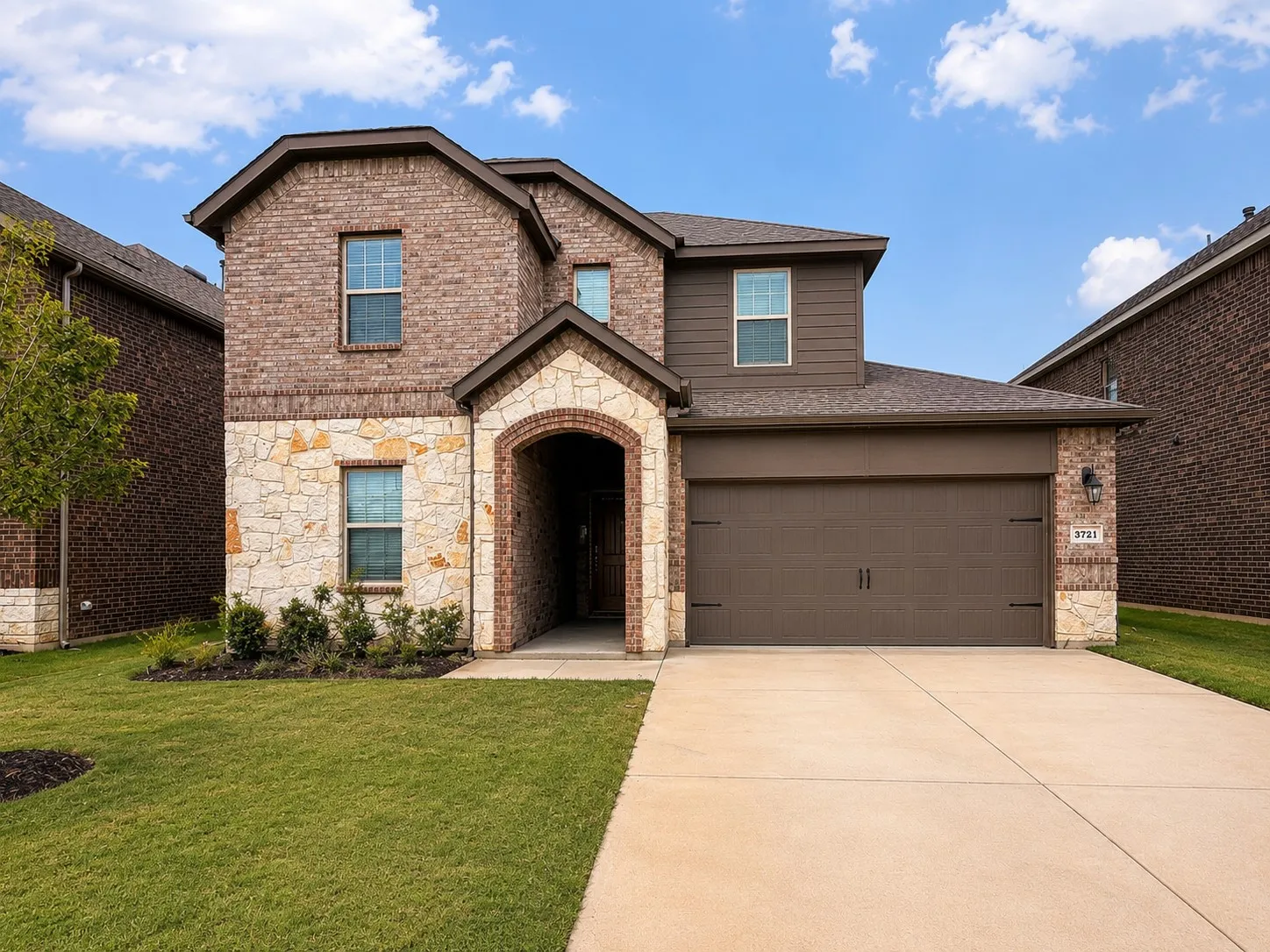 Brick and stone facade featuring an arched entryway, attached garage with carriage-style doors, concrete driveway, and established front lawn with landscaping