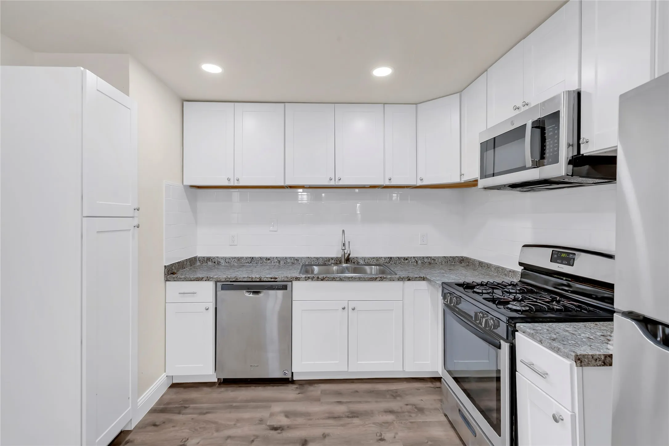 Kitchen featuring white shaker cabinetry, stainless steel appliances, speckled stone countertops, a white subway tile backsplash, and wood-finish flooring