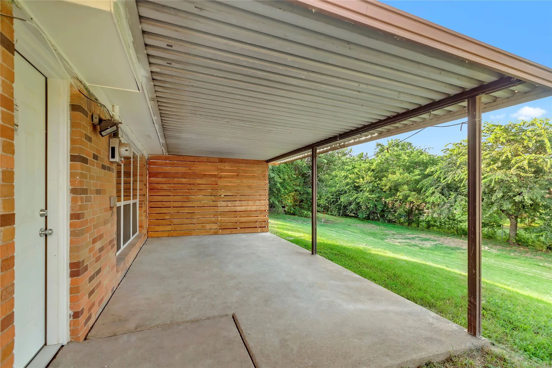 Expansive covered patio featuring a concrete slab, metal roofing, wood slat accent wall, and a brick exterior